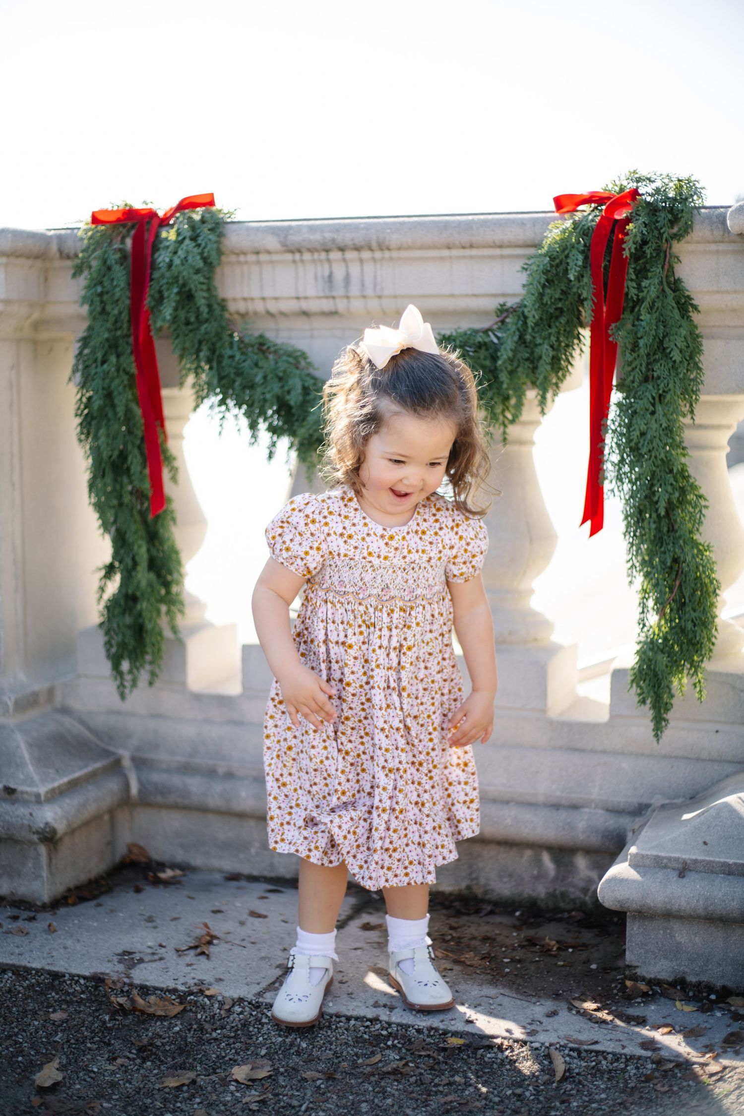 A child in a patterned dress explores a stone balcony decorated with Christmas garlands.