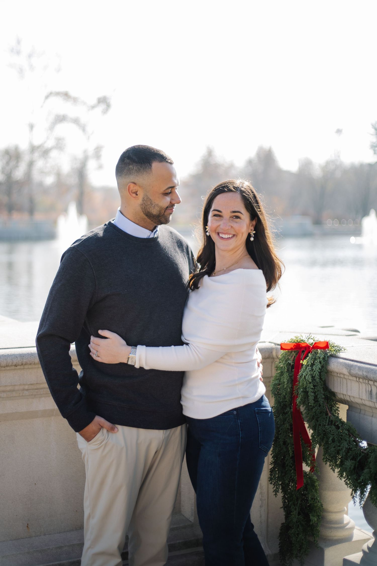 Couple embraces by lakeside with holiday greenery and red bow decorating stone railing.
