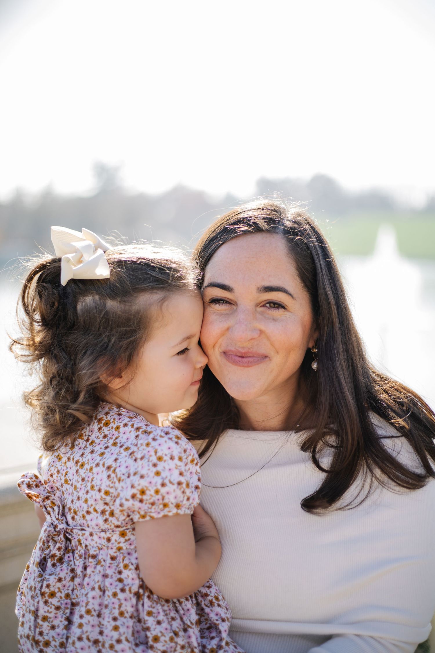 Sweet candid photos show a child in a floral dress giving kisses to their parent in an affectionate outdoor setting.
