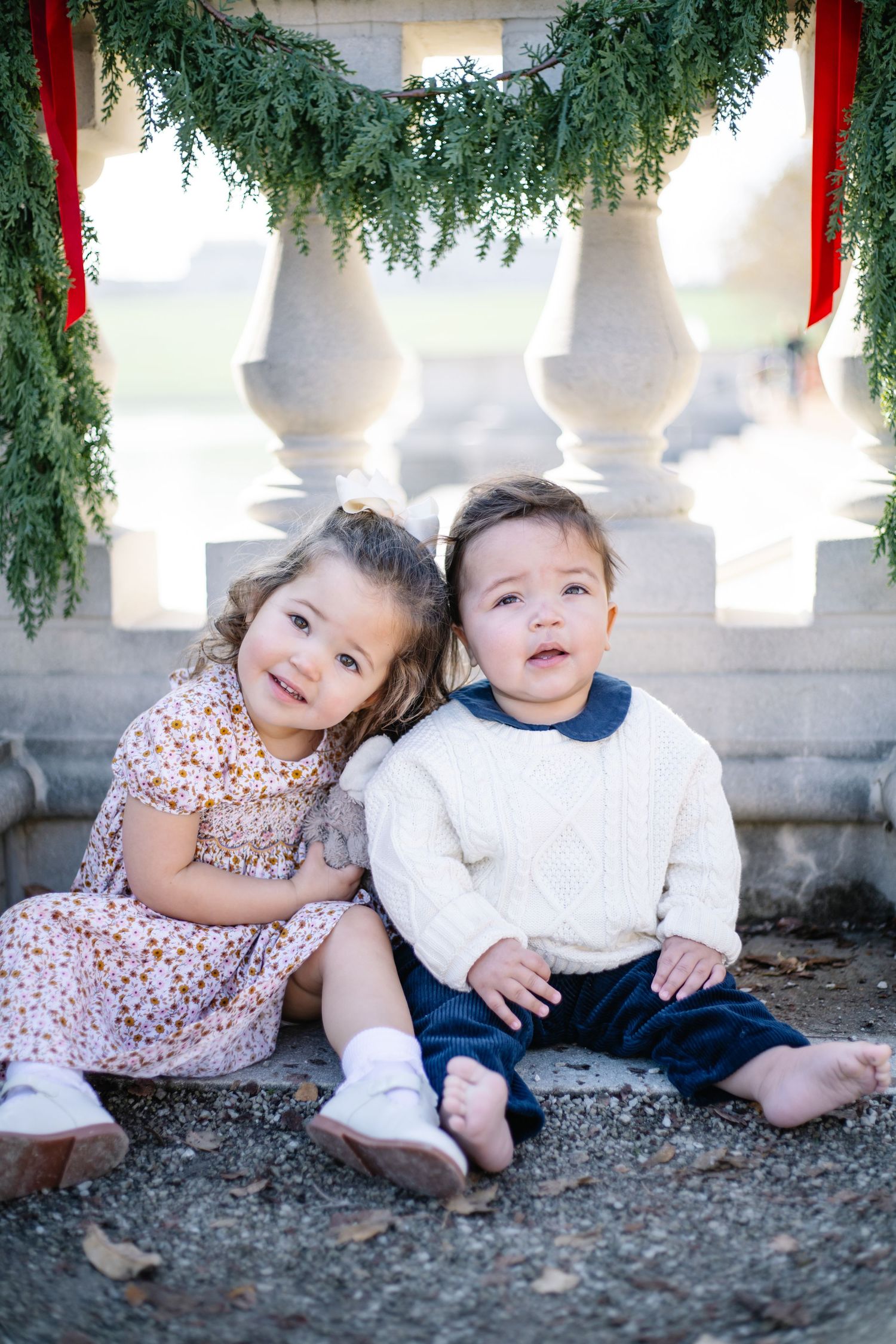 Two children sit together on stone steps decorated with holiday greenery and red ribbons.