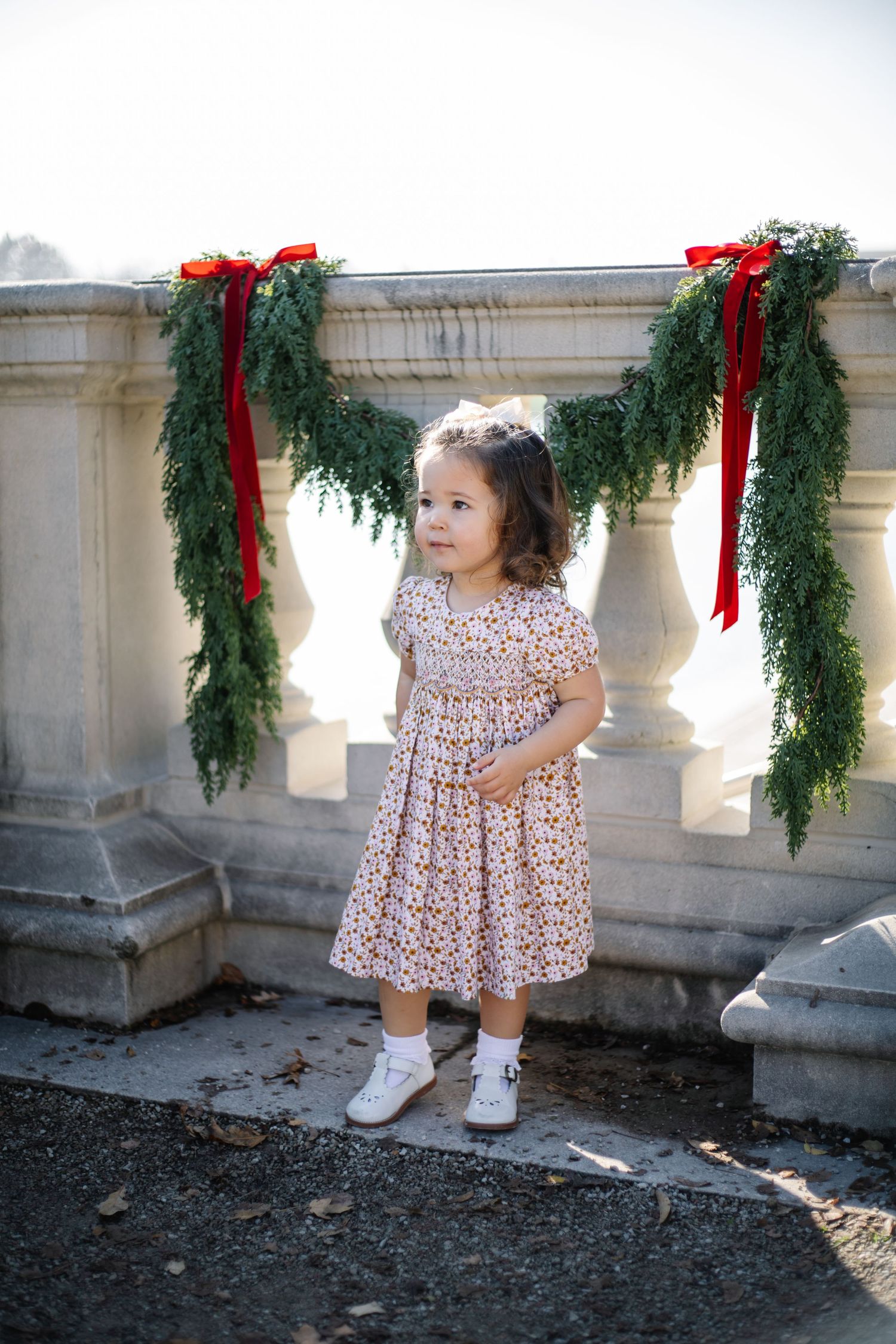 A toddler in a floral dress stands near stone columns decorated with holiday greenery and red bows.