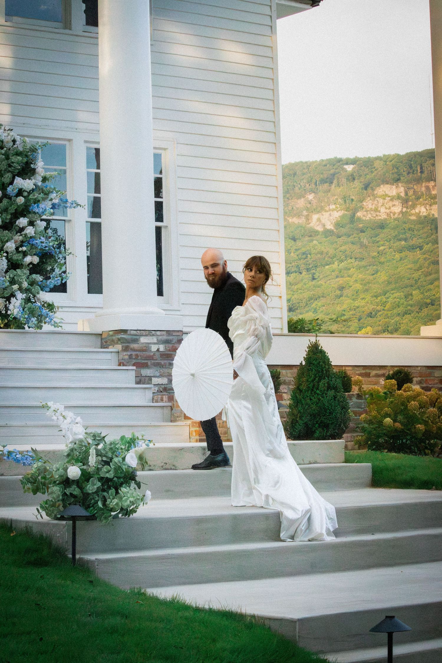 A couple shares a romantic moment on the steps of a white house with mountains in the background.