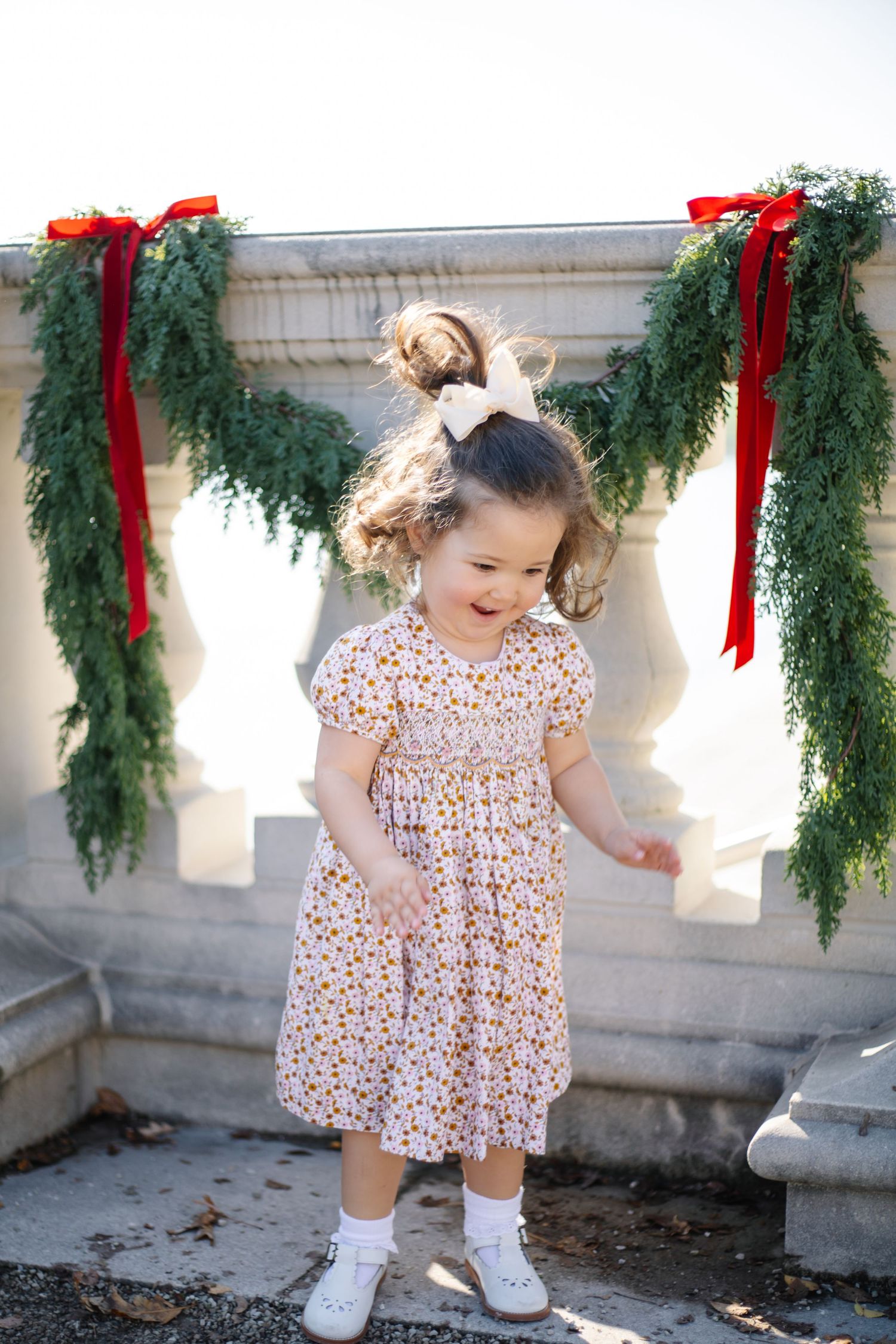 A toddler in a floral dress plays near holiday greenery with red bows.