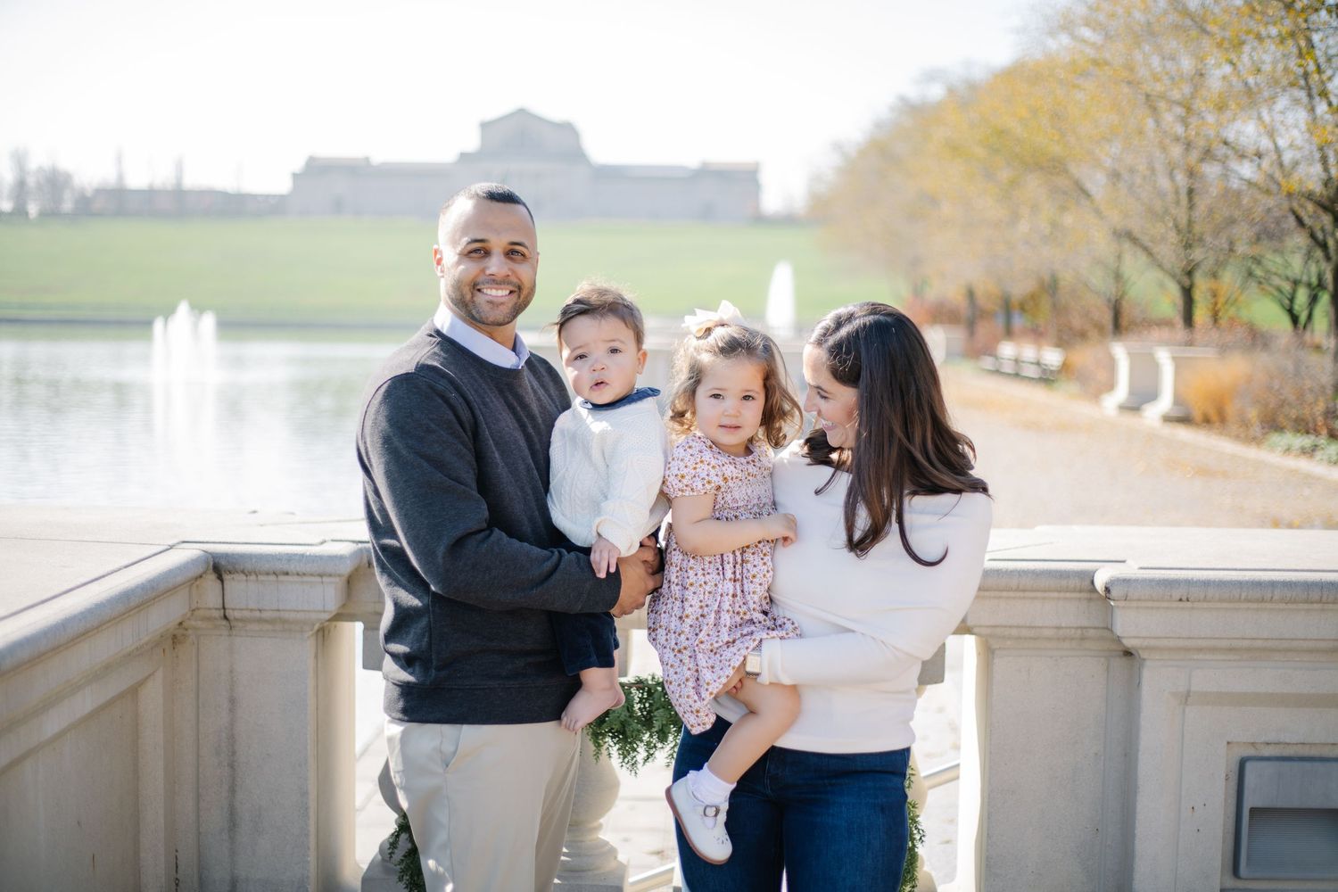 Family standing together on an outdoor stone balcony with trees and water in the background.