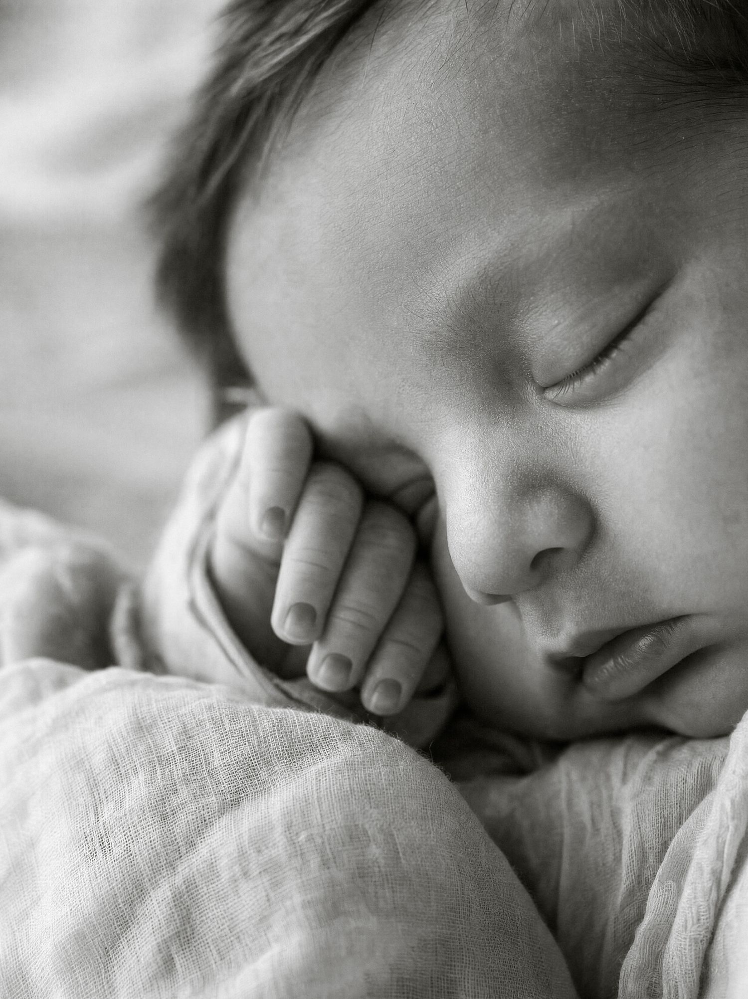 Black and white photo of a sleeping newborn baby with tiny hands tucked under cheek, wrapped in soft muslin cloth.