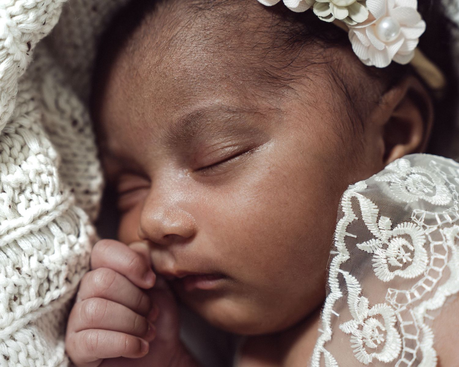 Sleeping newborn baby girl wearing a white floral headband, wrapped in delicate lace fabric.