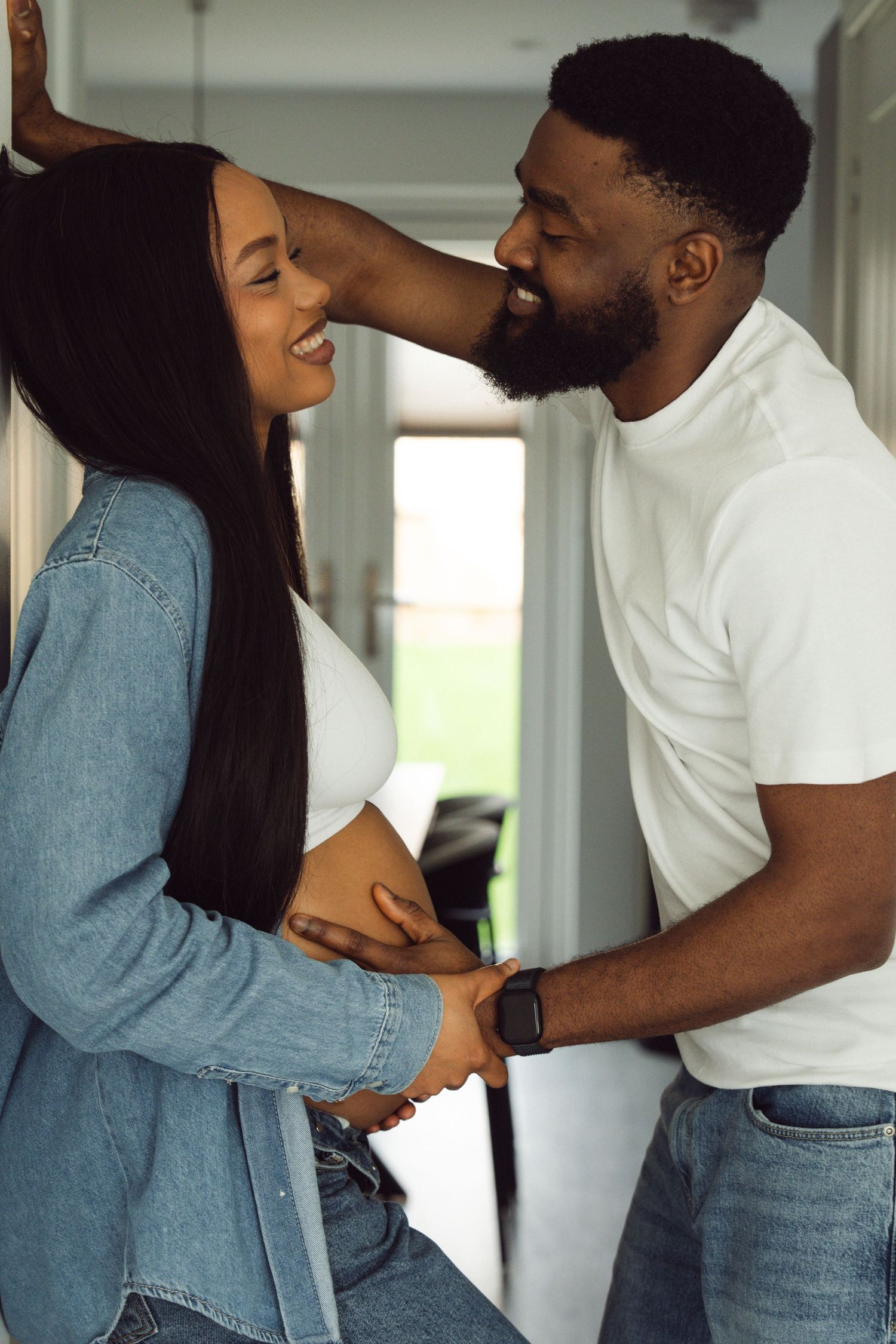 A happy pregnant Black couple smiling and holding baby bump together indoors, wearing casual denim outfits.