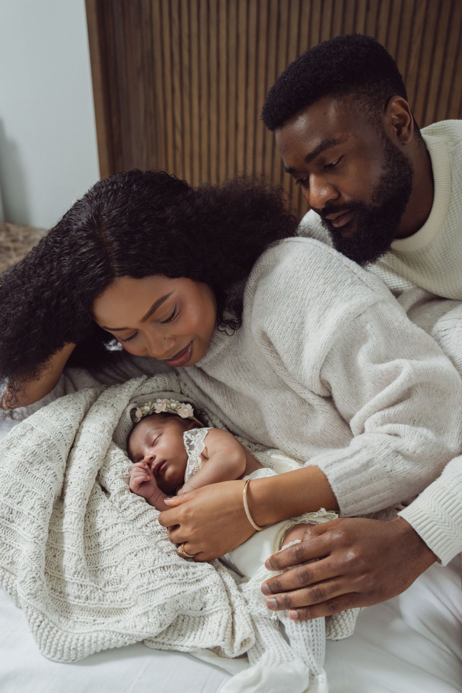 Black couple smiling at their sleeping newborn baby wrapped in a white knit blanket on a bed.