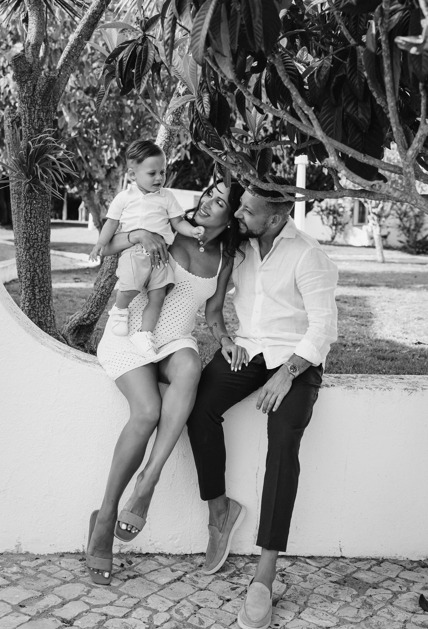 Family sitting together on white stone wall under palm trees.
