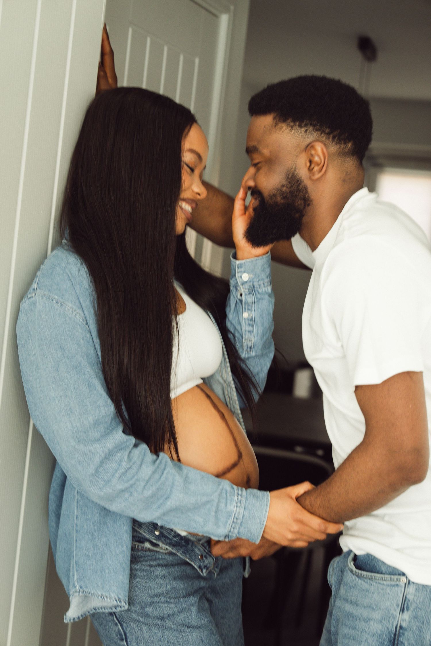 A pregnant Black woman in a denim jacket shares a tender moment with her partner in a cozy home setting.