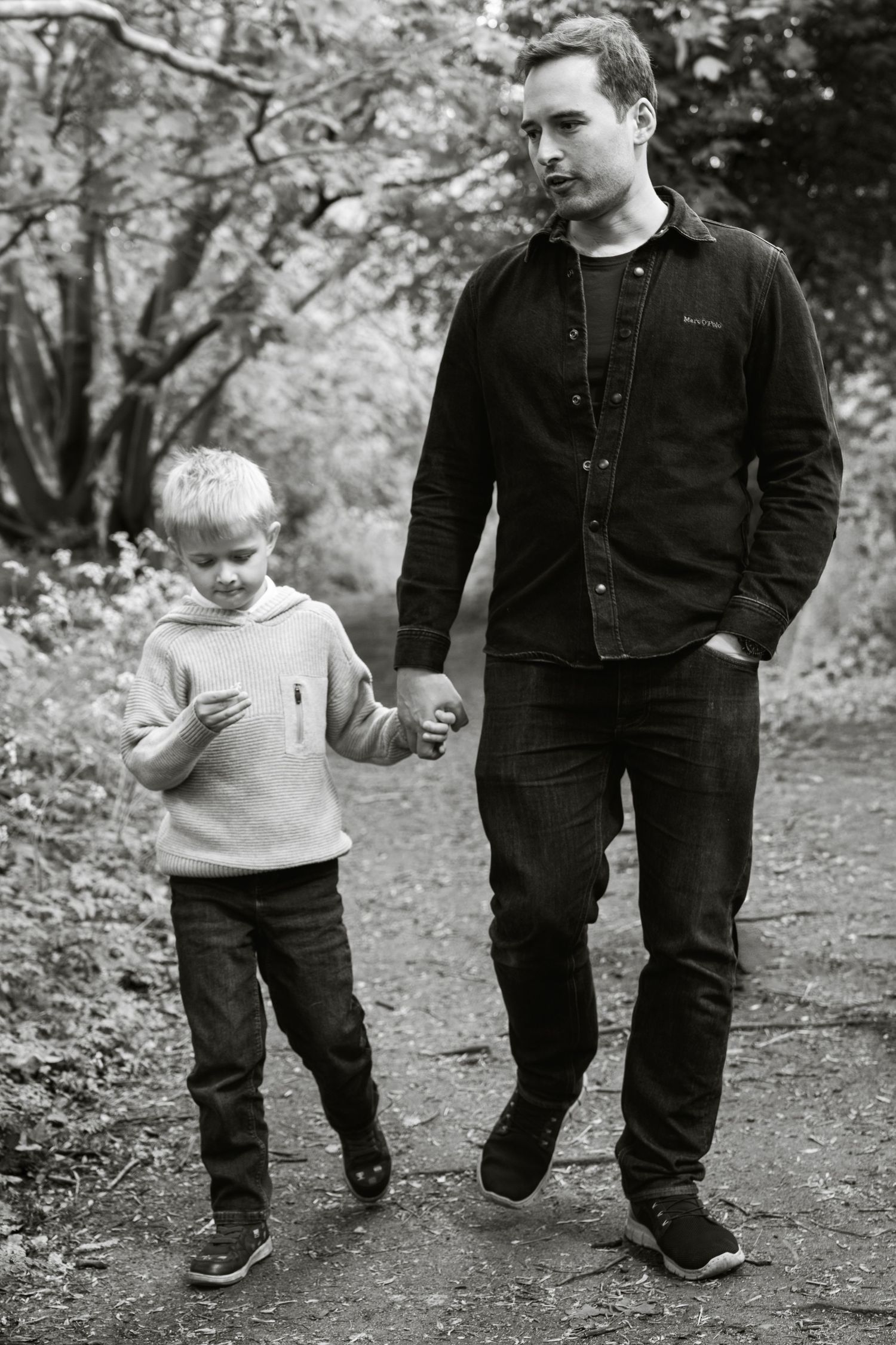 Black and white photo of a man and young boy holding hands while walking on a woodland path.