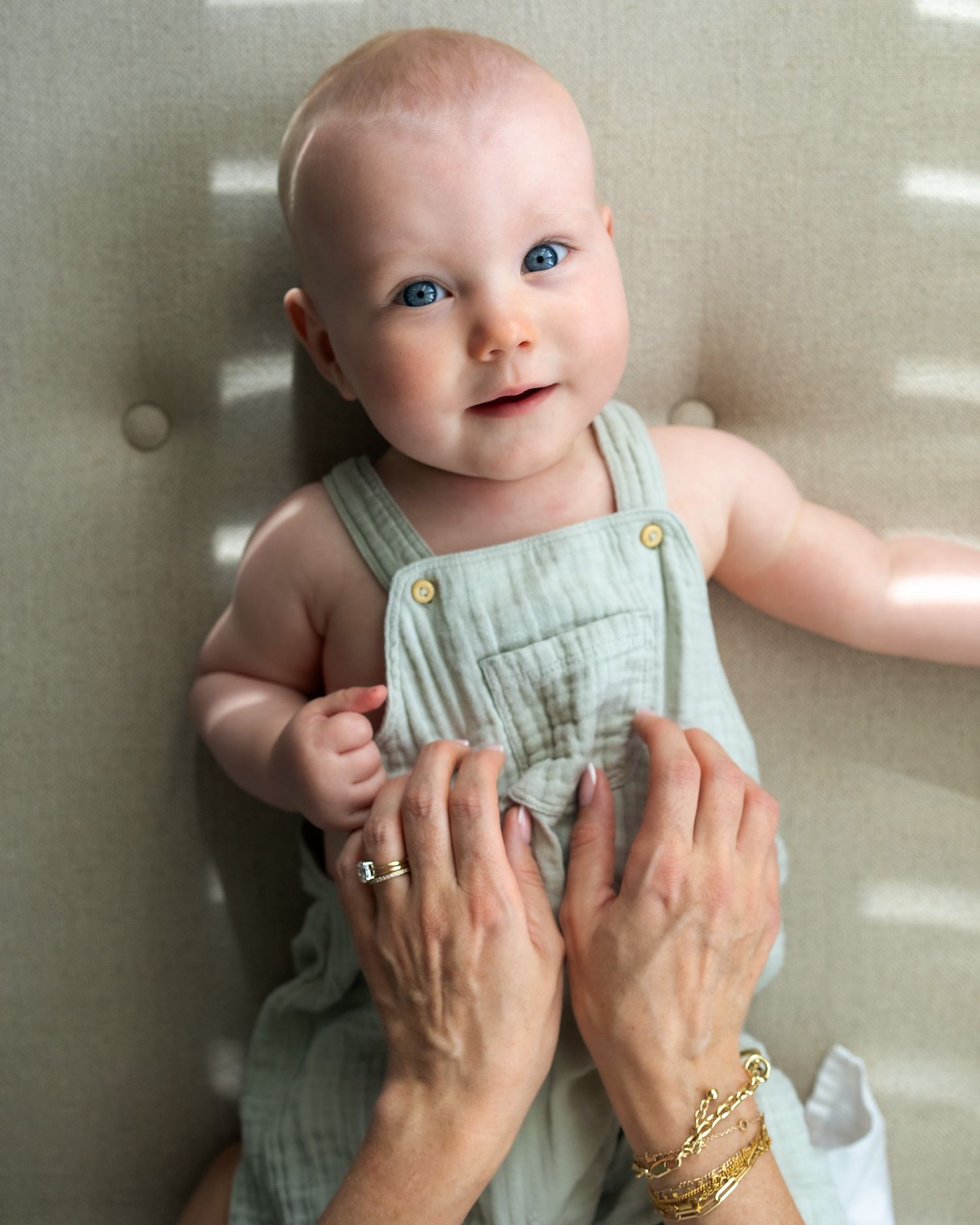 Baby in mint green overalls smiling while supported by adult hands.