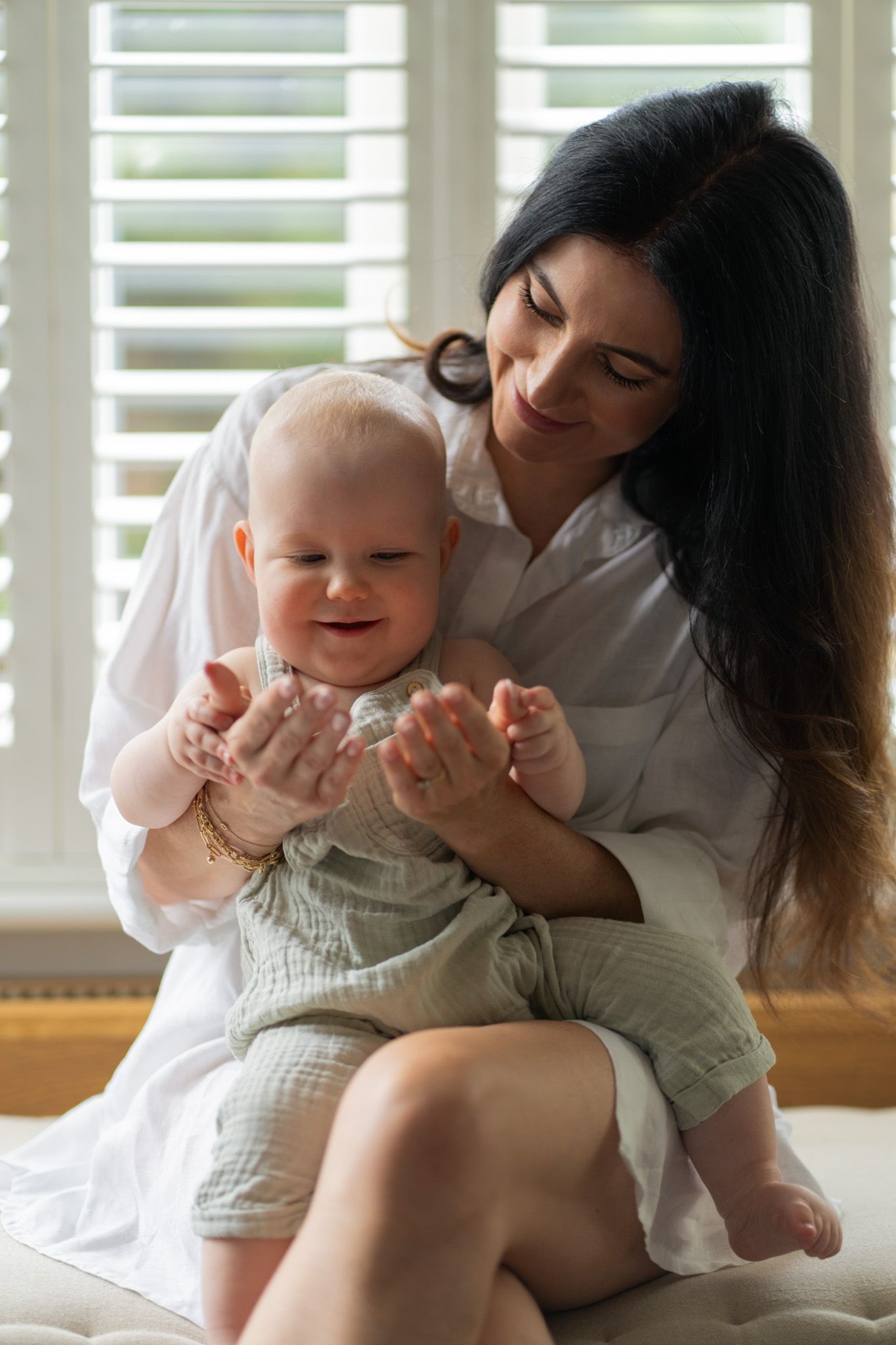 A tender indoor moment showing a parent and baby sharing joyful cuddles on a couch near window shutters.