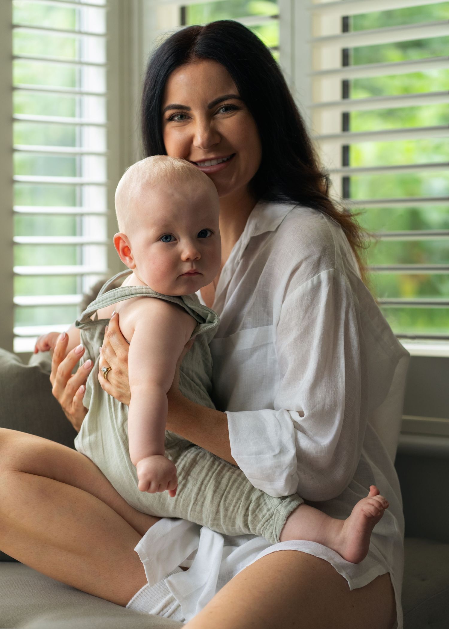 Tender moment between parent and baby by bright window with plantation shutters.