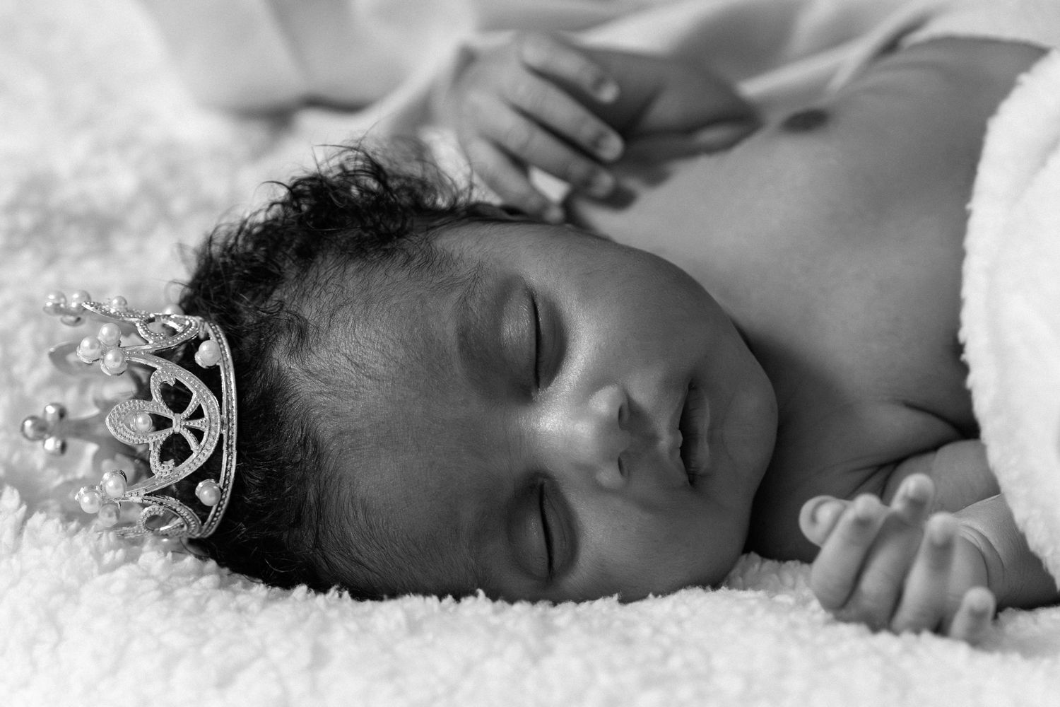 Sleeping newborn baby wearing a tiny tiara crown, black and white portrait photography on soft white blanket.
