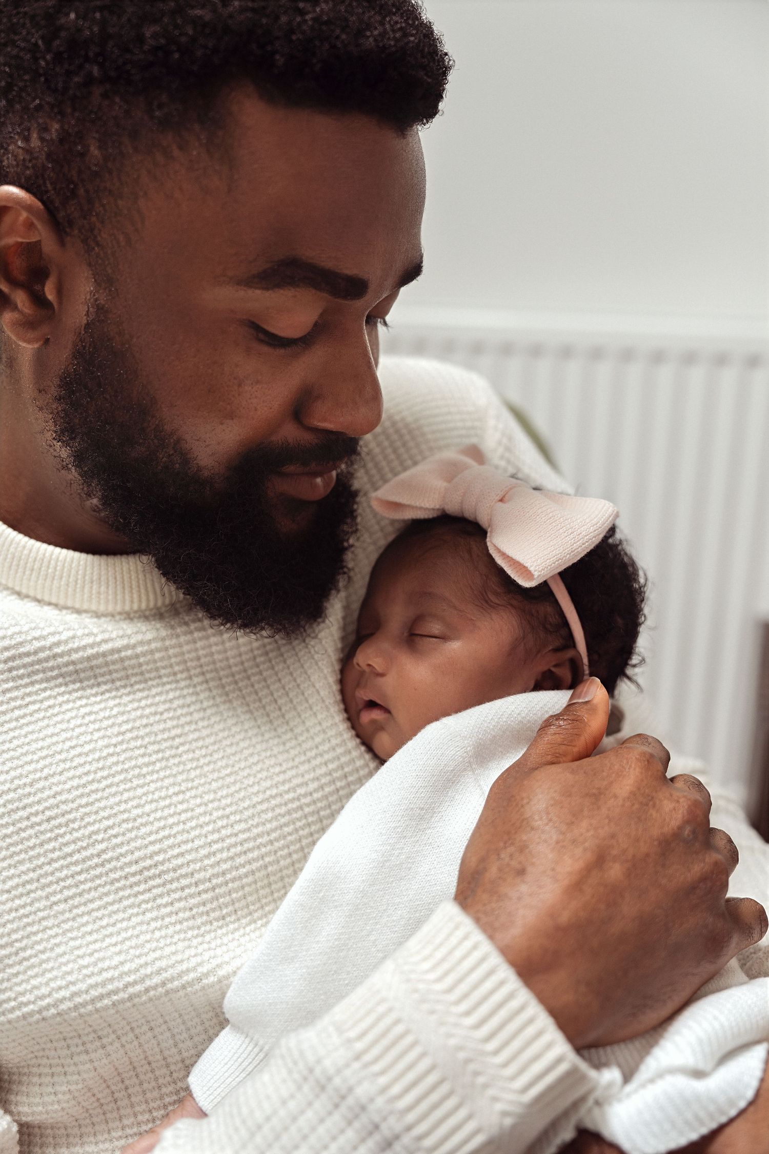 A bearded father tenderly holds his sleeping newborn baby girl wearing a pink bow headband.