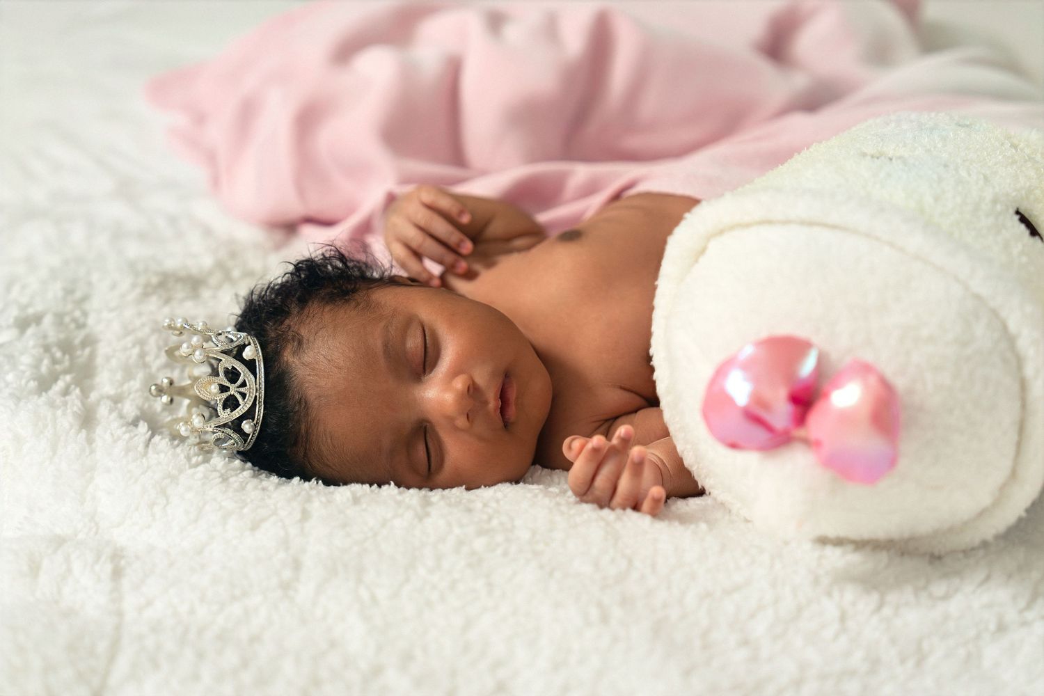 Newborn baby sleeping peacefully wearing a tiny tiara crown next to a white stuffed bunny on a fluffy white blanket.