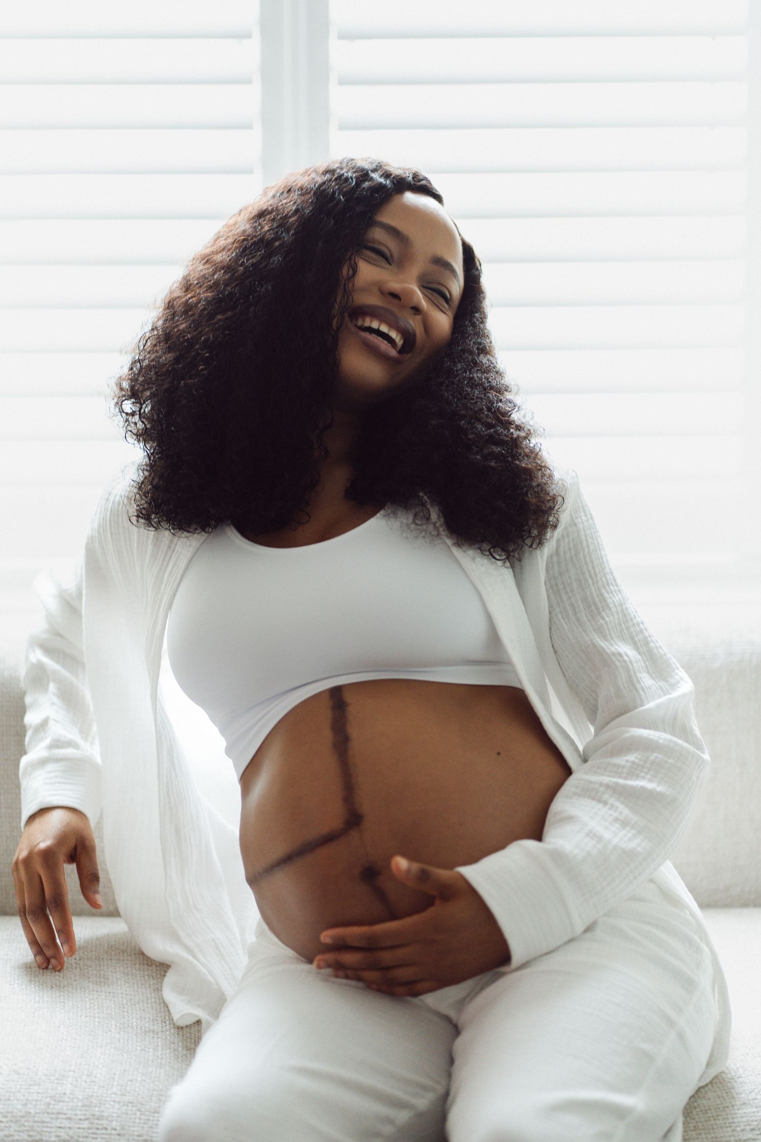 Smiling pregnant Black woman in white outfit cradling her baby bump while sitting on a couch near bright windows.