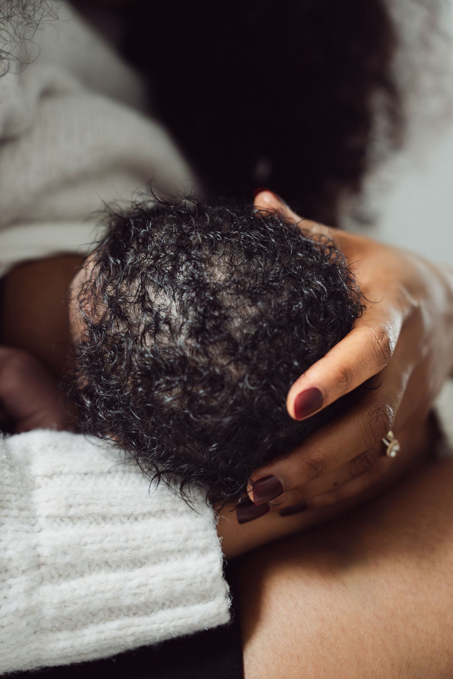 Close-up of a mother cradling her newborn baby's curly hair head with red manicured nails and gold ring.