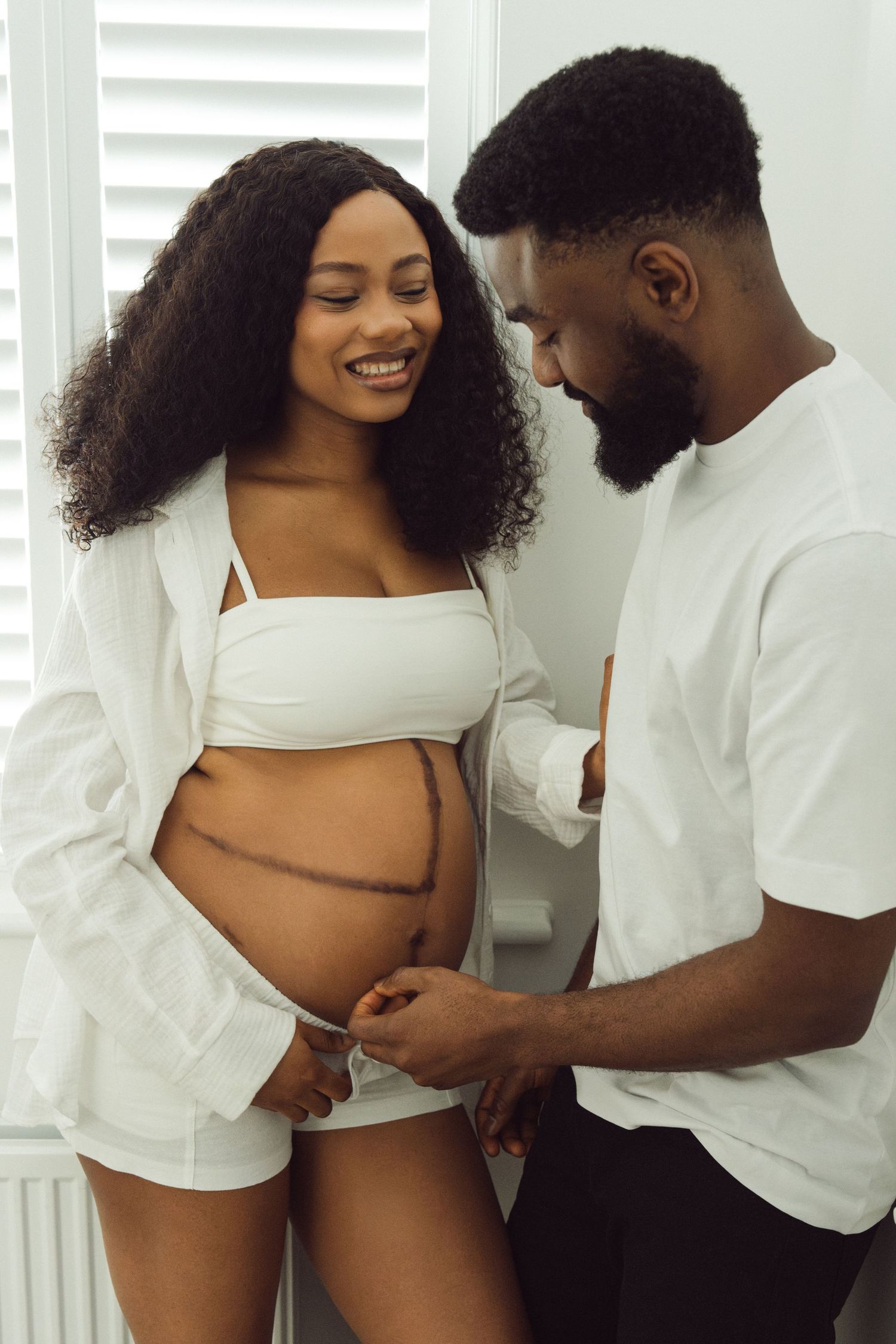 Expectant couple in white outfits, partner's hand on baby bump indoors | At-home maternity photographer in London by Inga Franlund