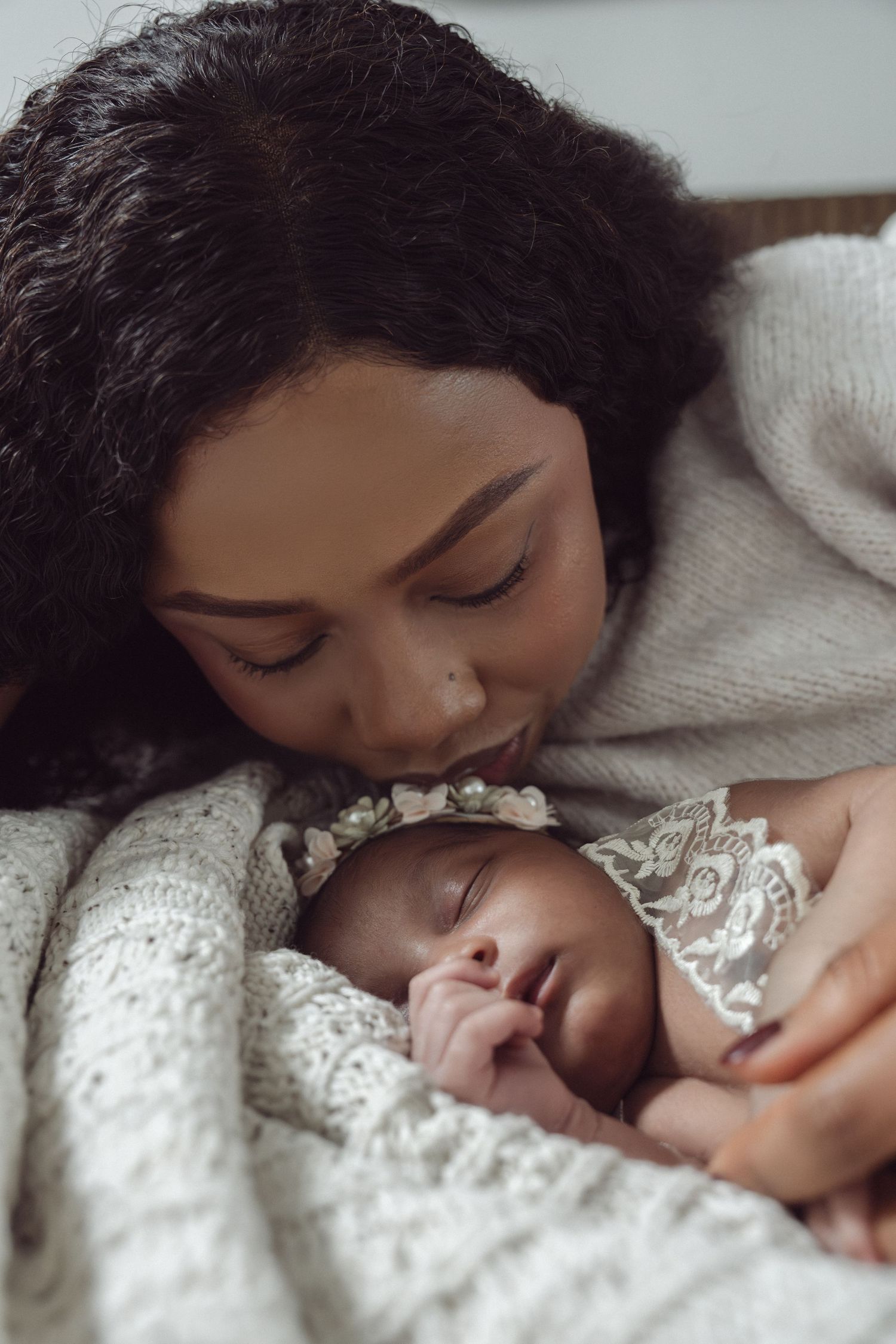A mother tenderly kisses her sleeping newborn baby girl wearing a floral headband and lace outfit wrapped in a white knit blanket.