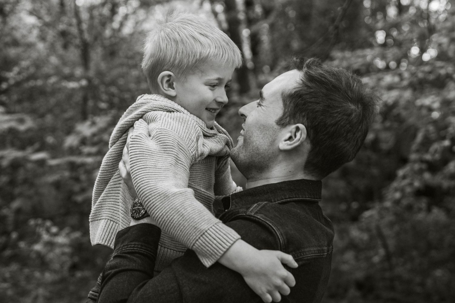 Black and white photo of a father lifting his young smiling son outdoors in a lush green forest setting.