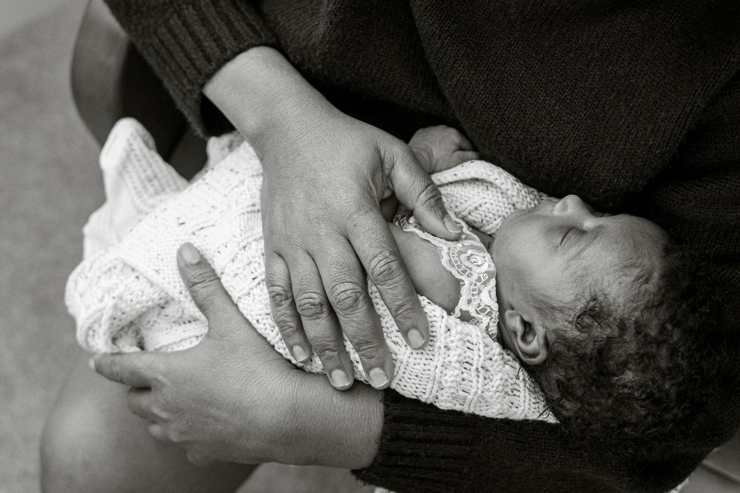 Black and white photo of a newborn baby cradled in loving hands, wrapped in a delicate white knit blanket.