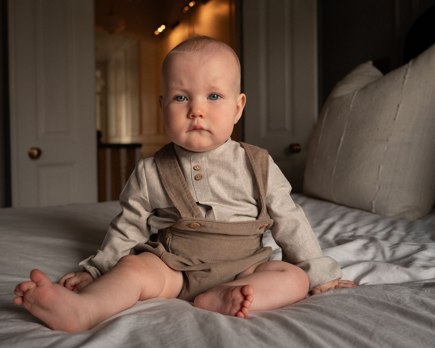 A baby wearing a tan collared outfit sits on white bedding in a series of progressive photos.