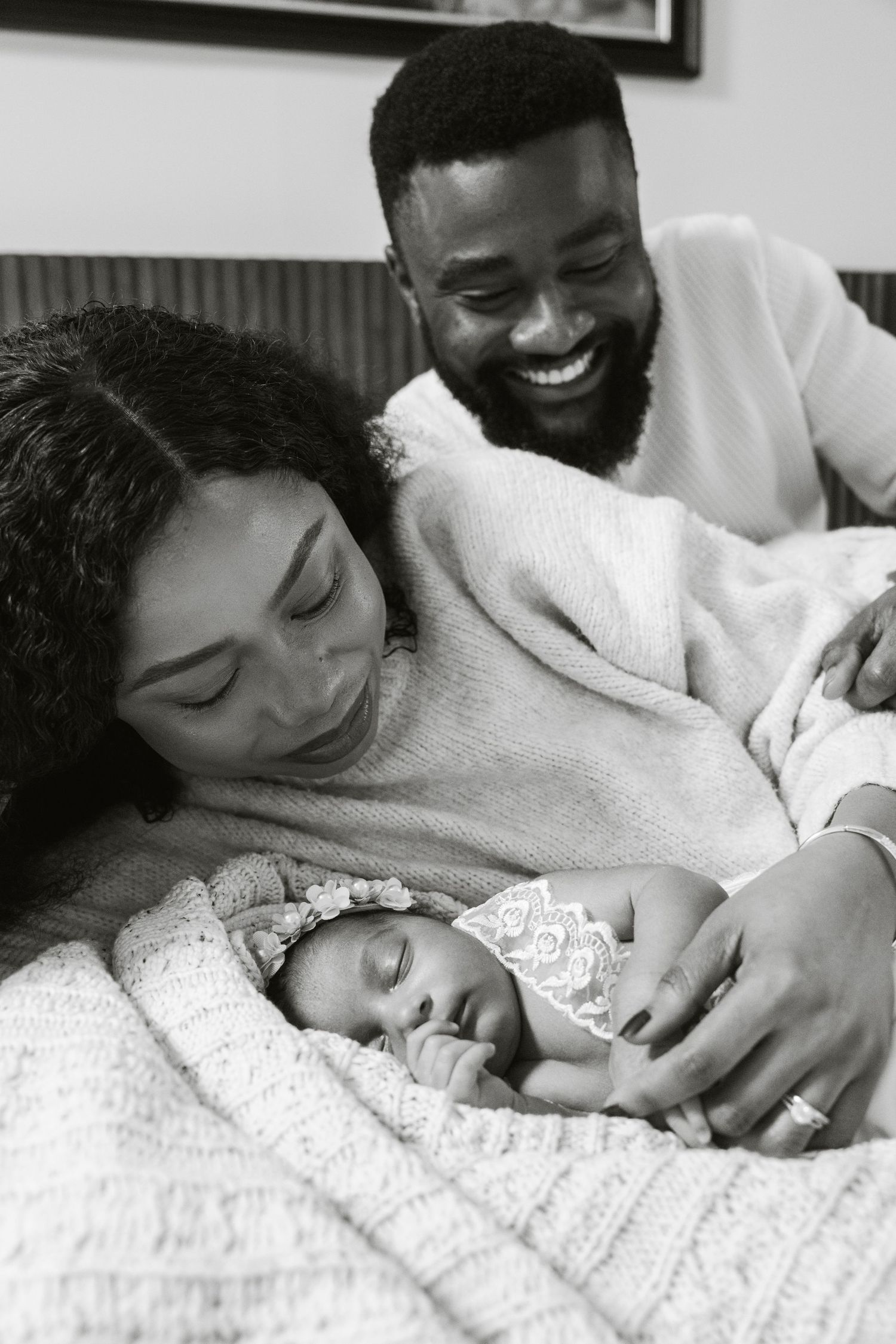 Black and white photo of smiling parents lovingly gazing at their sleeping newborn baby wrapped in a blanket.