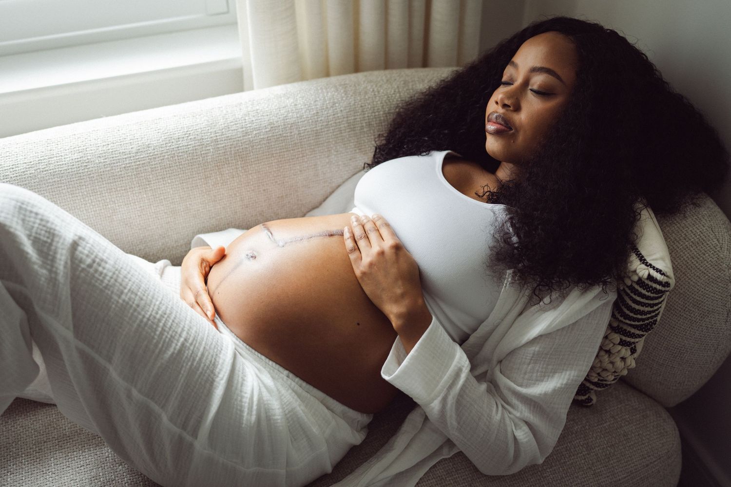 Pregnant Black woman relaxing on sofa, cradling baby bump, wearing white outfit, with curly hair and eyes closed.