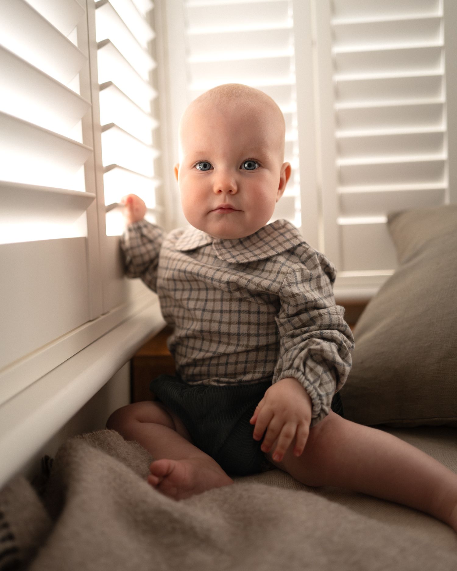 A baby in a plaid shirt sits by a window with white shutters in a series of candid moments.