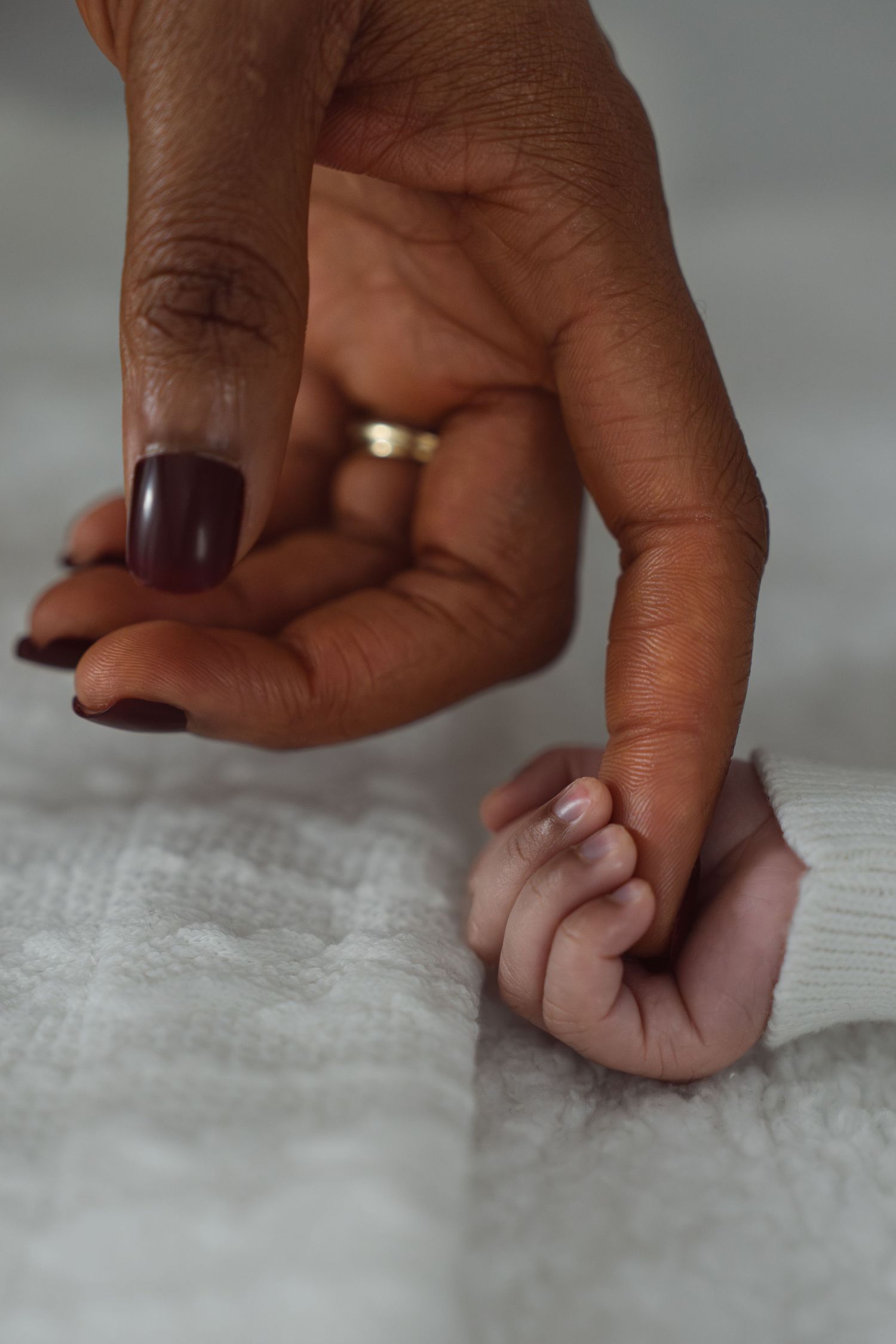 A newborn baby grasping the finger of a mother with dark nail polish and a gold wedding ring.