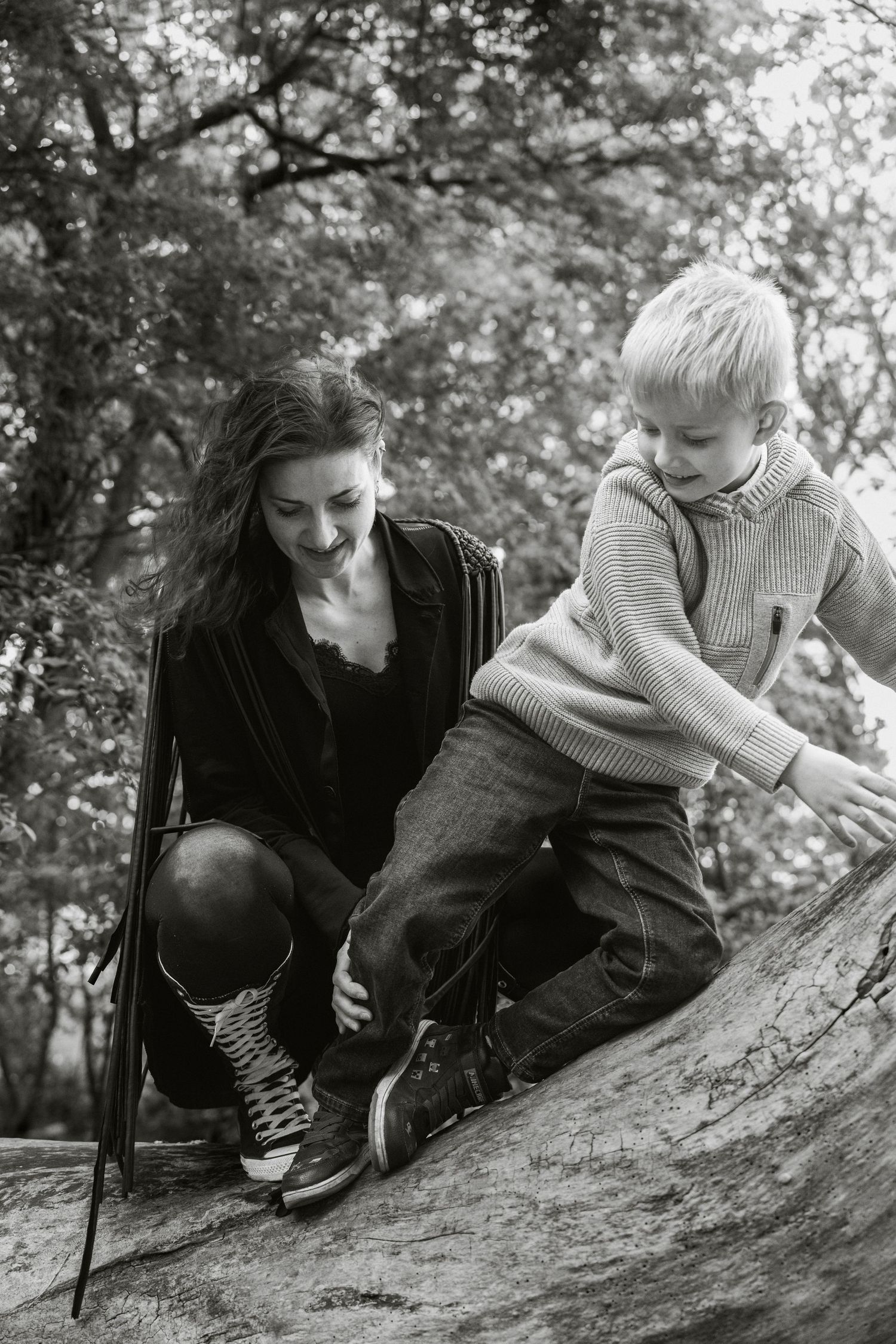 Black and white photo of a smiling woman helping a young blonde boy climb a large rock in a forest.