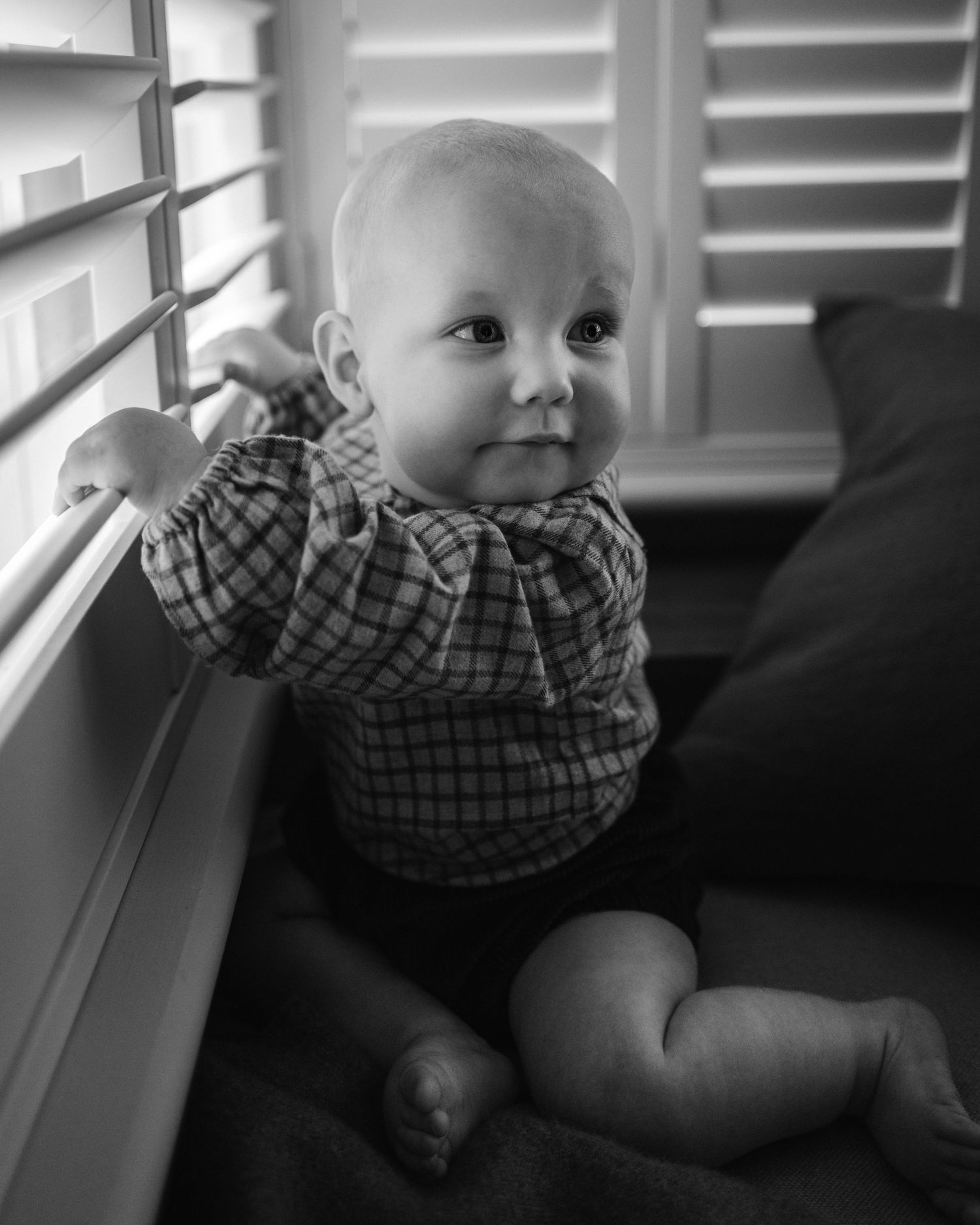 Baby in plaid shirt looking curiously through window blinds.