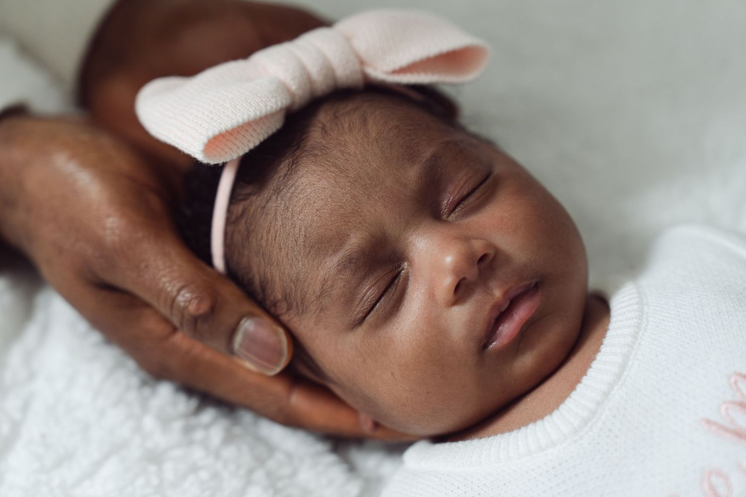Sleeping newborn baby girl wearing a pink bow headband, cradled gently in an adult's hands.
