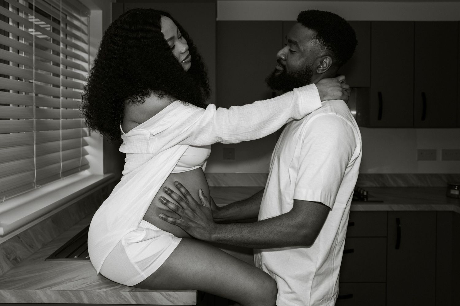Black and white photo of expectant couple in kitchen, woman seated on counter | At-home maternity photographer in London by Inga Franlund