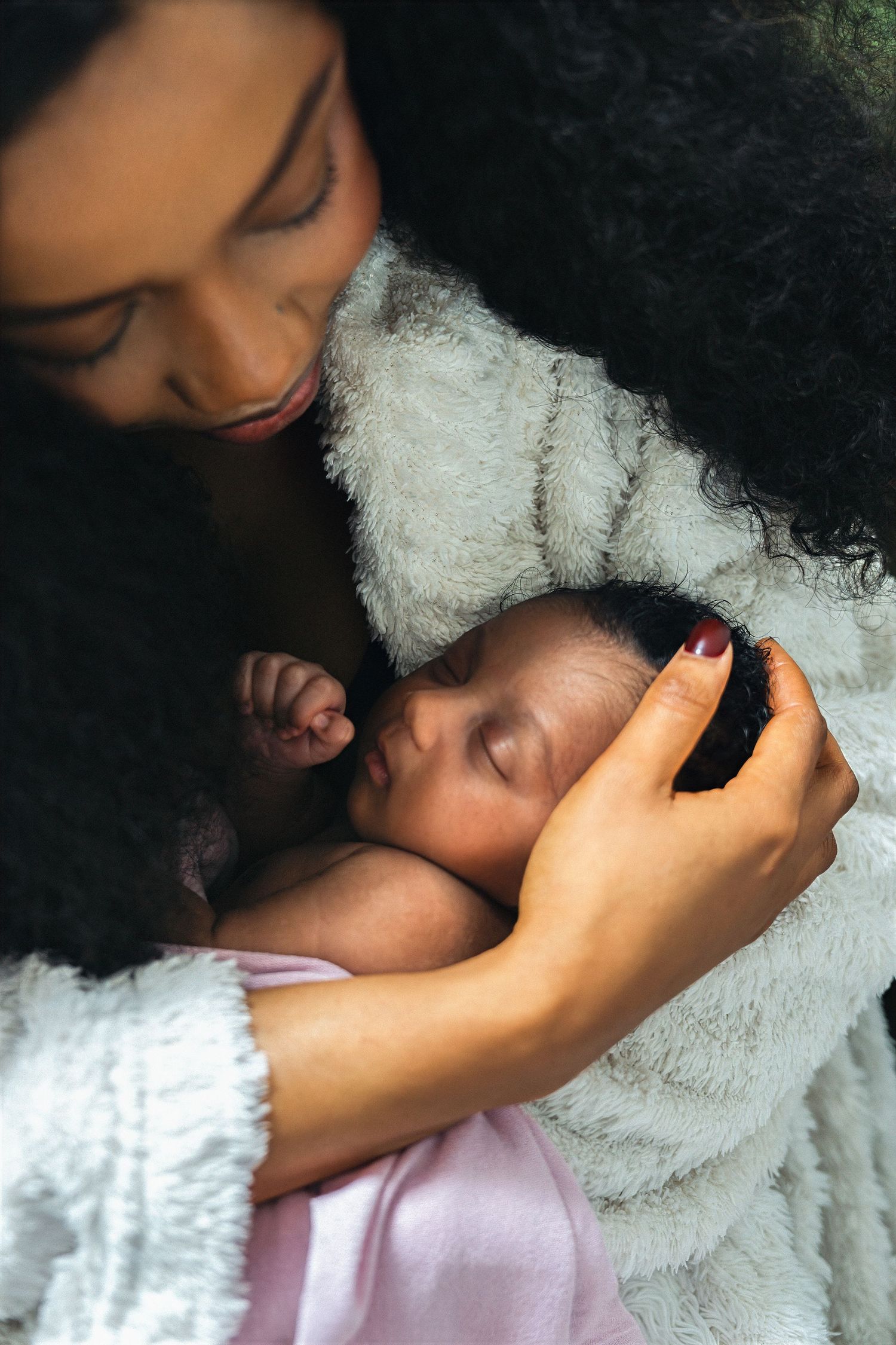 A mother with curly hair tenderly holds her sleeping newborn baby wrapped in a soft pink blanket.