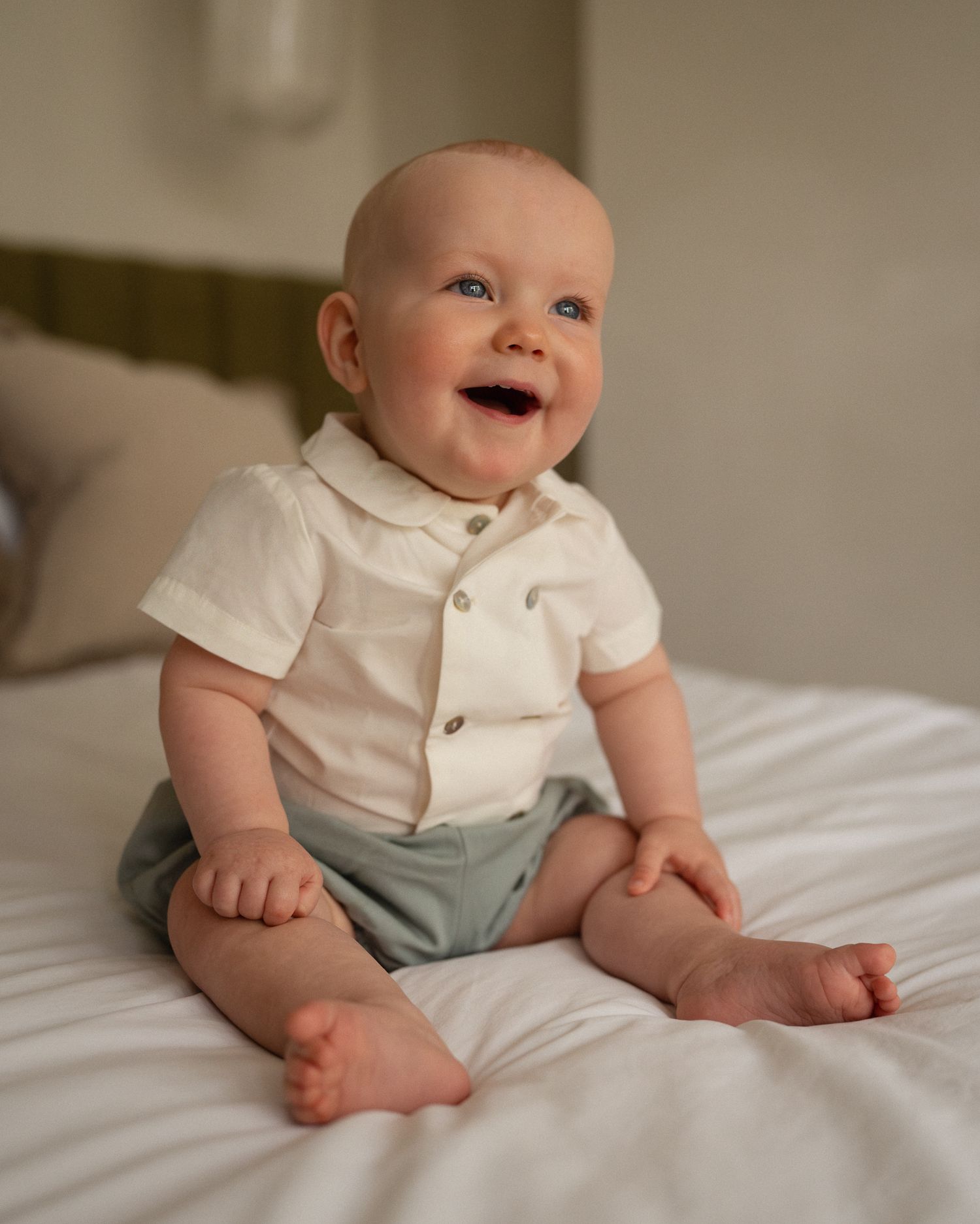 A baby in a white collared shirt and blue pants sits on white bedding showing various expressions and poses.