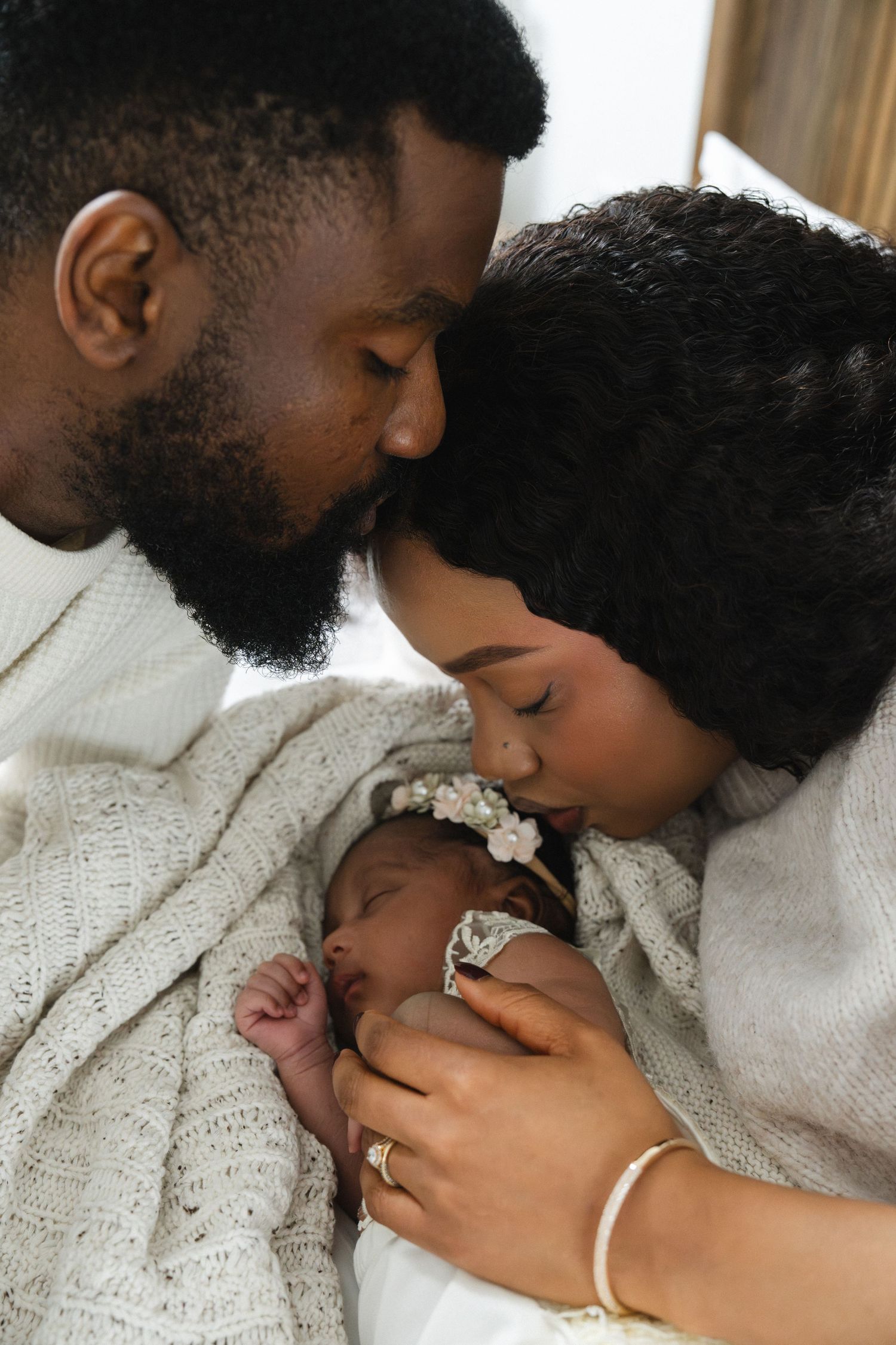 Black parents lovingly kiss their sleeping newborn baby wrapped in white during an intimate family portrait.