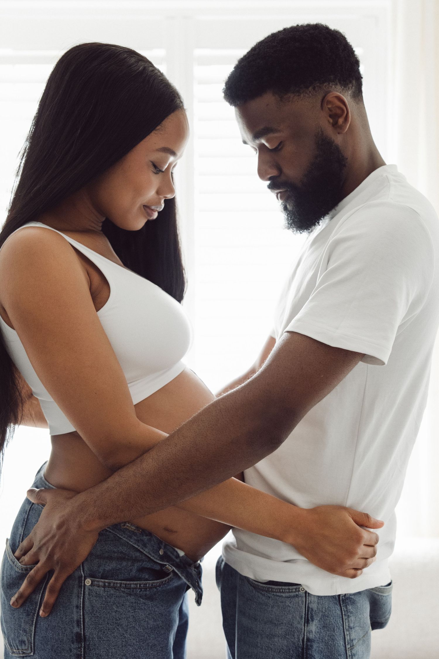 Expectant couple tenderly looking down at baby bump, hands together | At-home maternity photographer in London by Inga Franlund