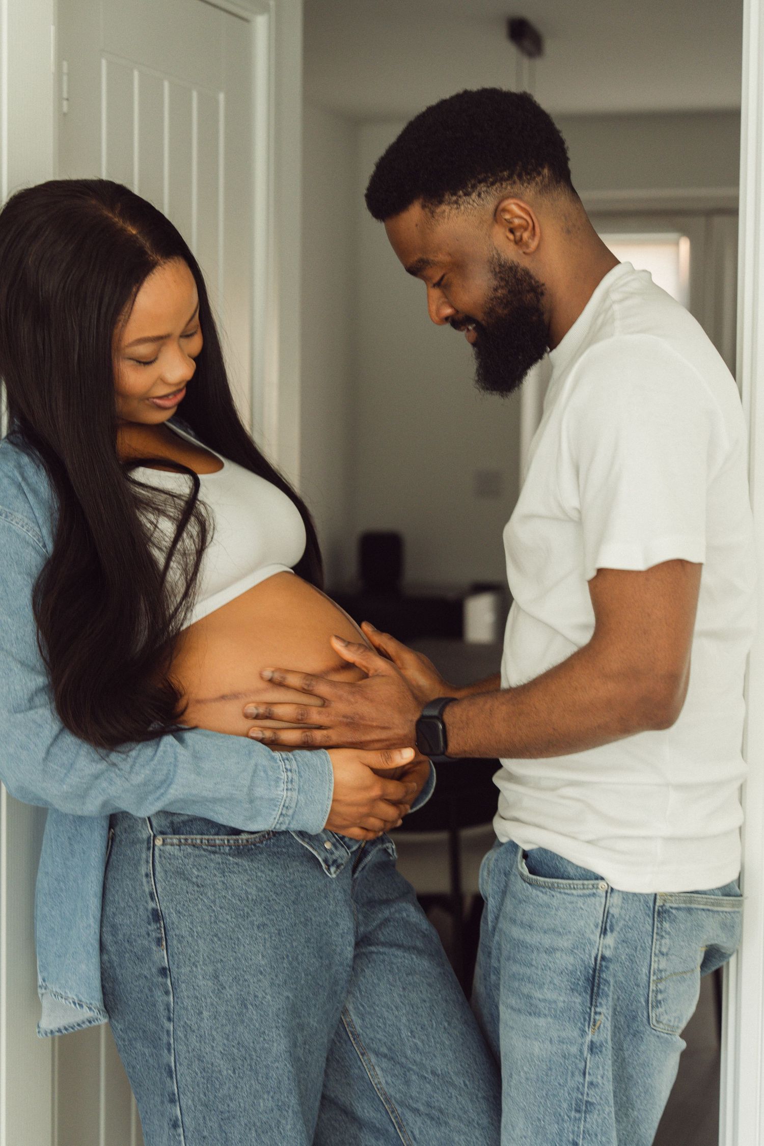 Expectant couple in jeans and white tops, hands cradling baby bump | At-home maternity photographer in London by Inga Franlund