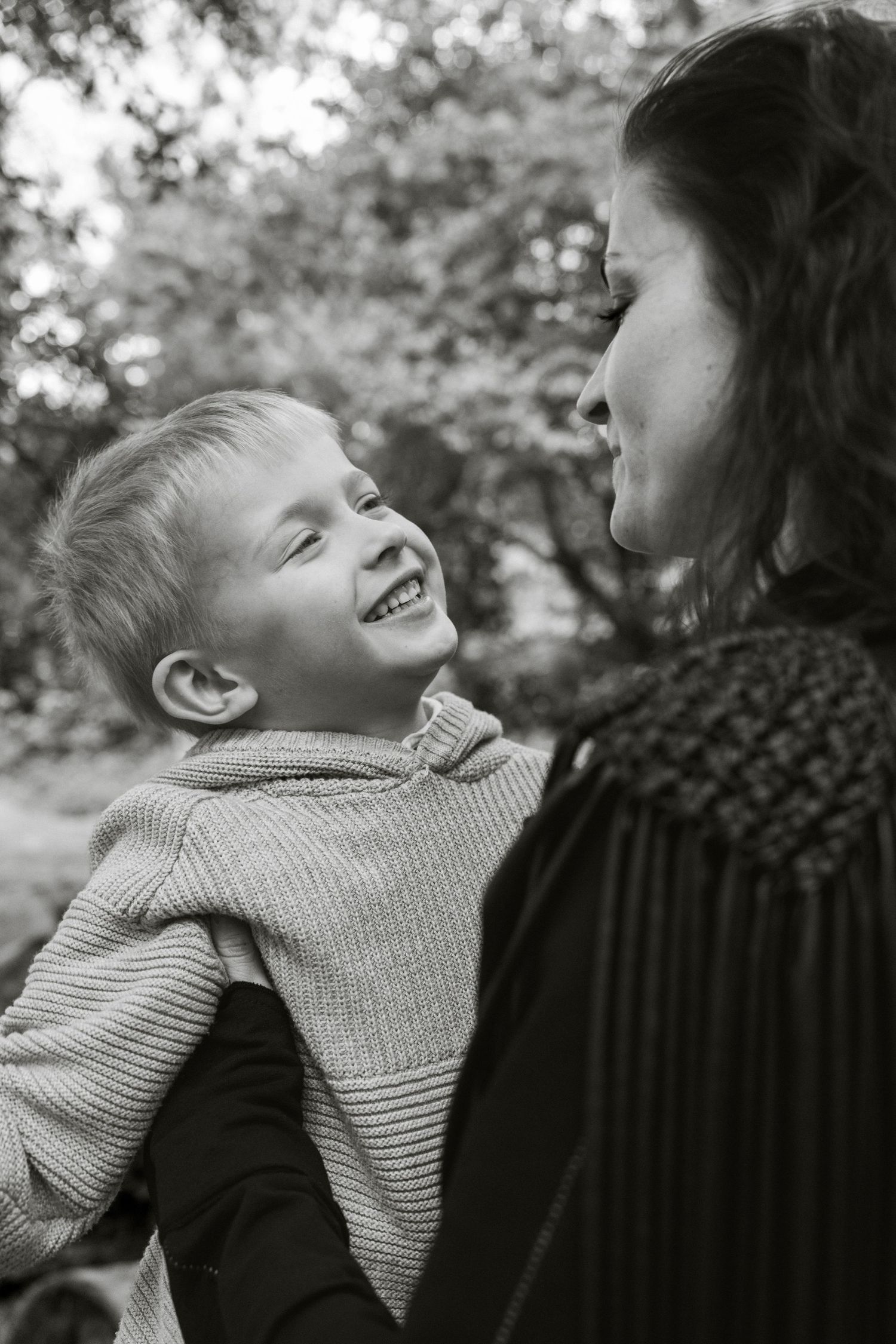 Black and white photo of a smiling boy being held by a woman outdoors among trees.