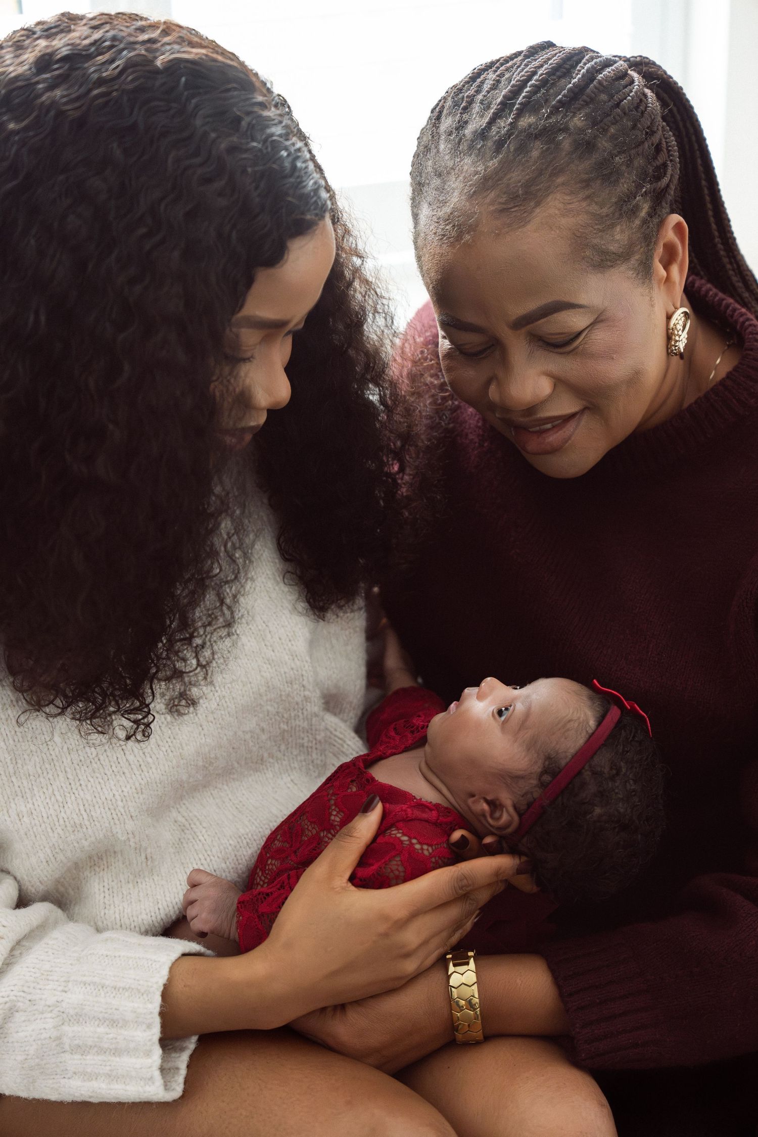Two women lovingly gaze at a newborn baby dressed in red, sharing a tender three-generation family moment.