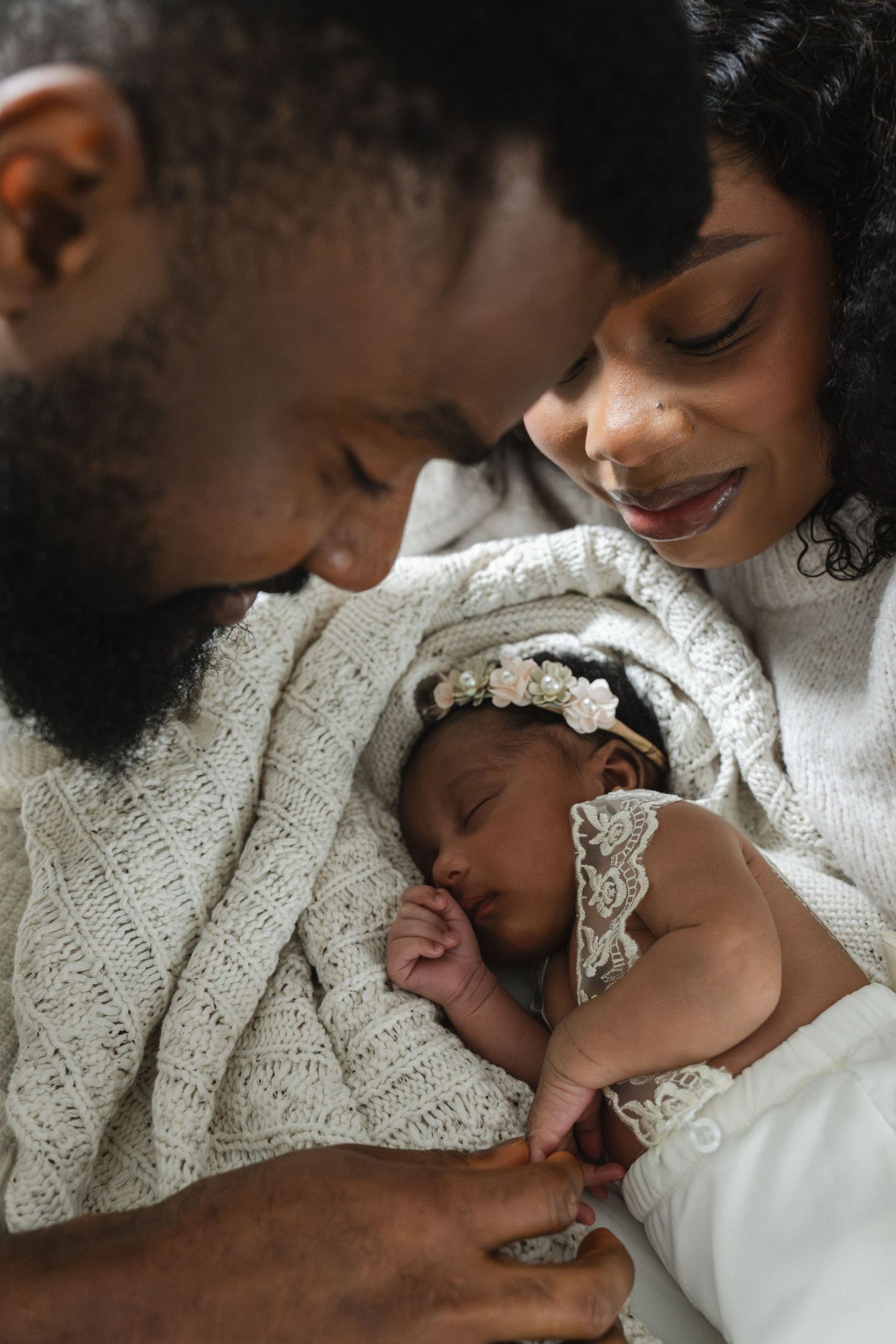 Loving parents smiling at their sleeping newborn baby girl wrapped in a white knit blanket.