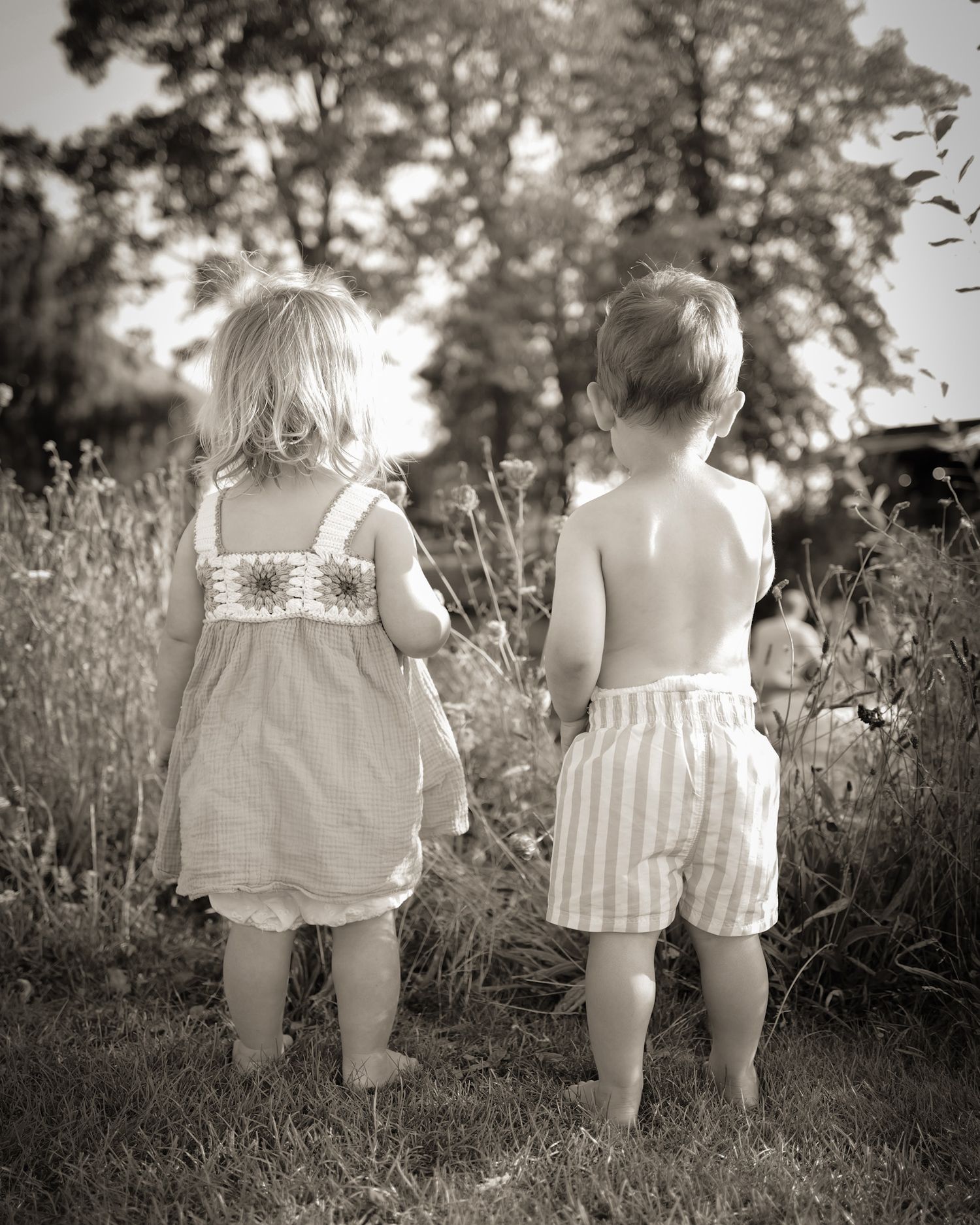 Two small children in summer outfits walking together through a grassy field in black and white.