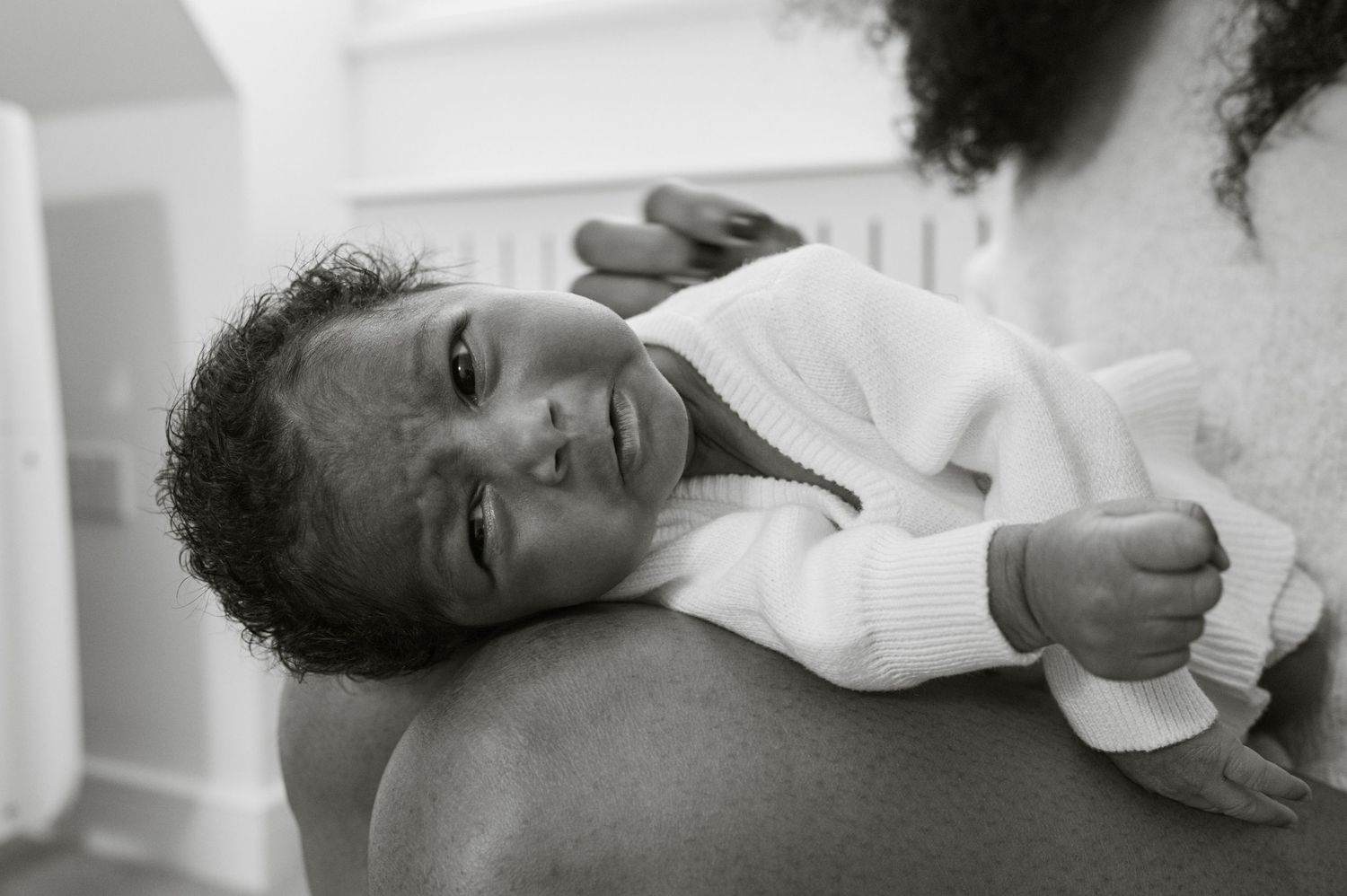 Black and white photo of a newborn baby resting on an adult's shoulder, wearing a white knit sweater.