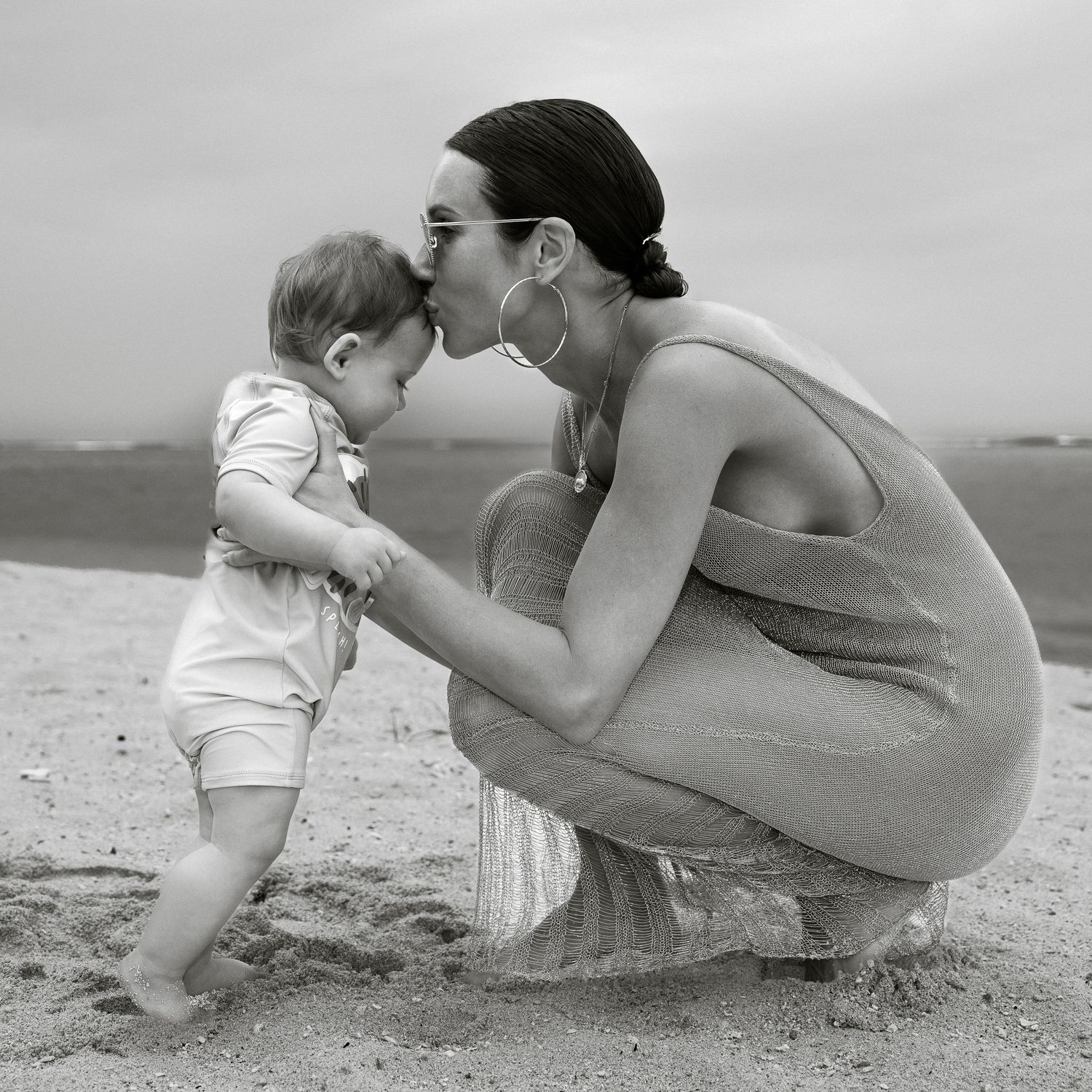 Black and white photo of a mother crouching on a beach, kissing her baby while holding hands near the shoreline.