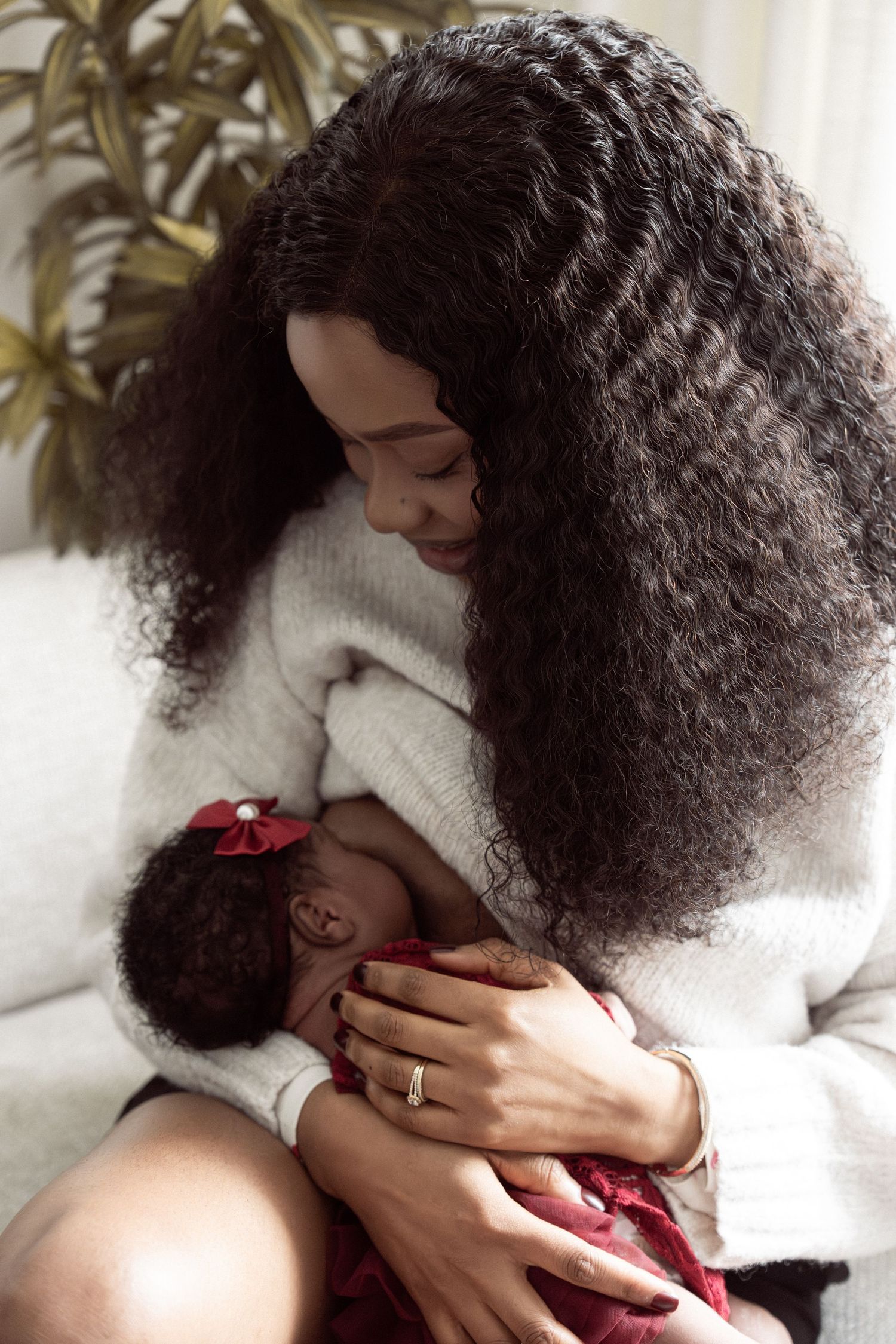 A Black mother with long curly hair tenderly holds her newborn baby girl in a red outfit while sitting on a couch.
