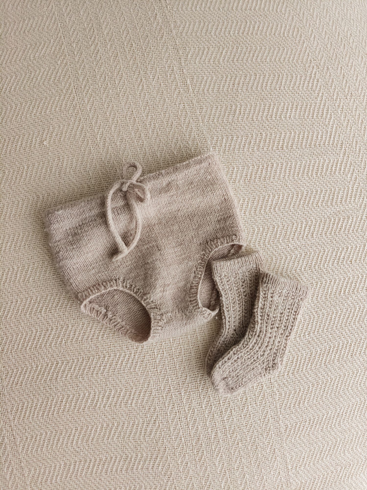 Handknit beige baby bloomers and matching crochet socks set in natural oatmeal yarn on textured background.