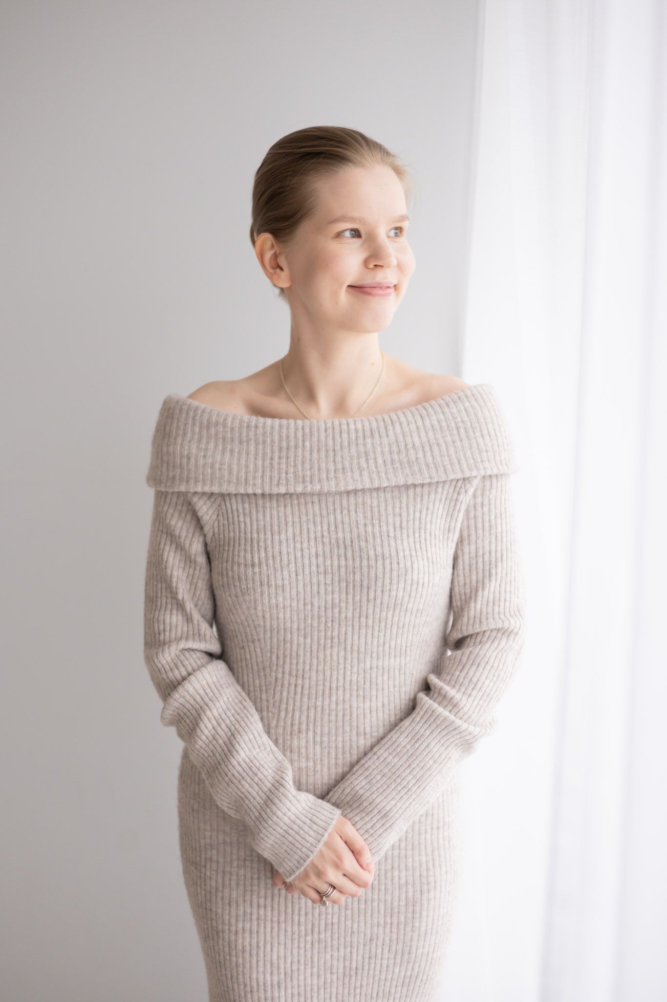 A person wearing a cozy off-shoulder ribbed gray sweater dress stands near a white window with a gentle smile.